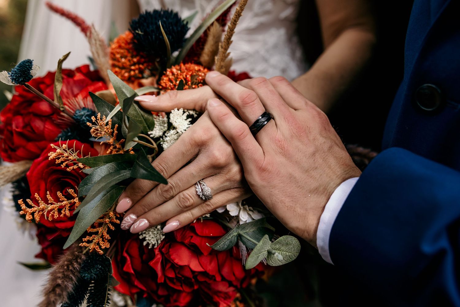 Close-up of wedding rings and hands holding a vibrant red floral bouquet with blue accents.