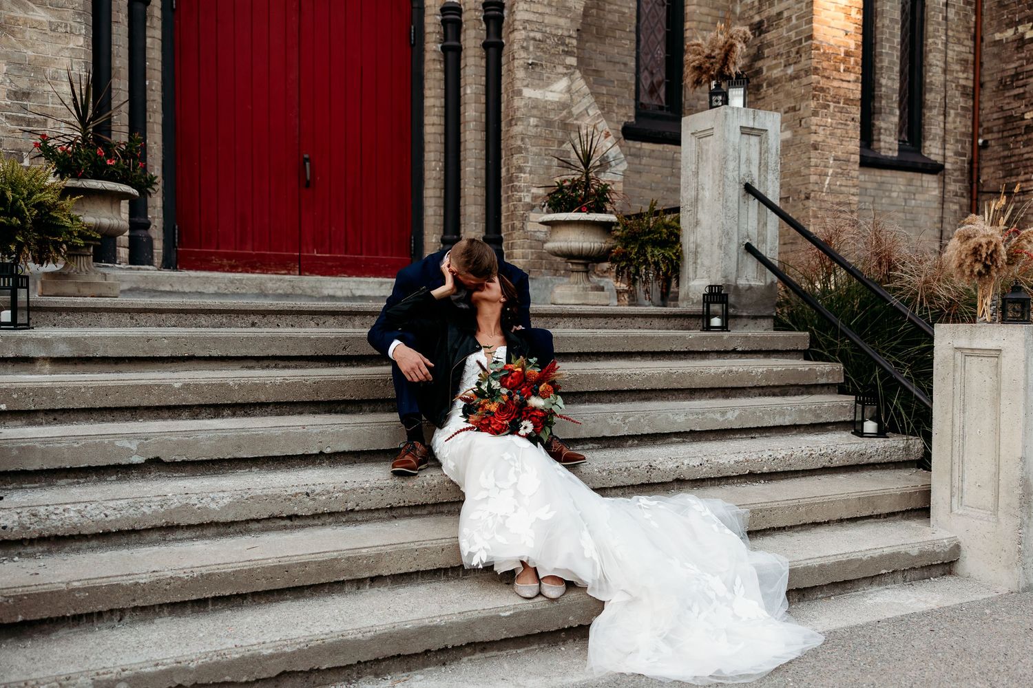 A wedding couple sits on stone steps in front of a historic building with a red door.