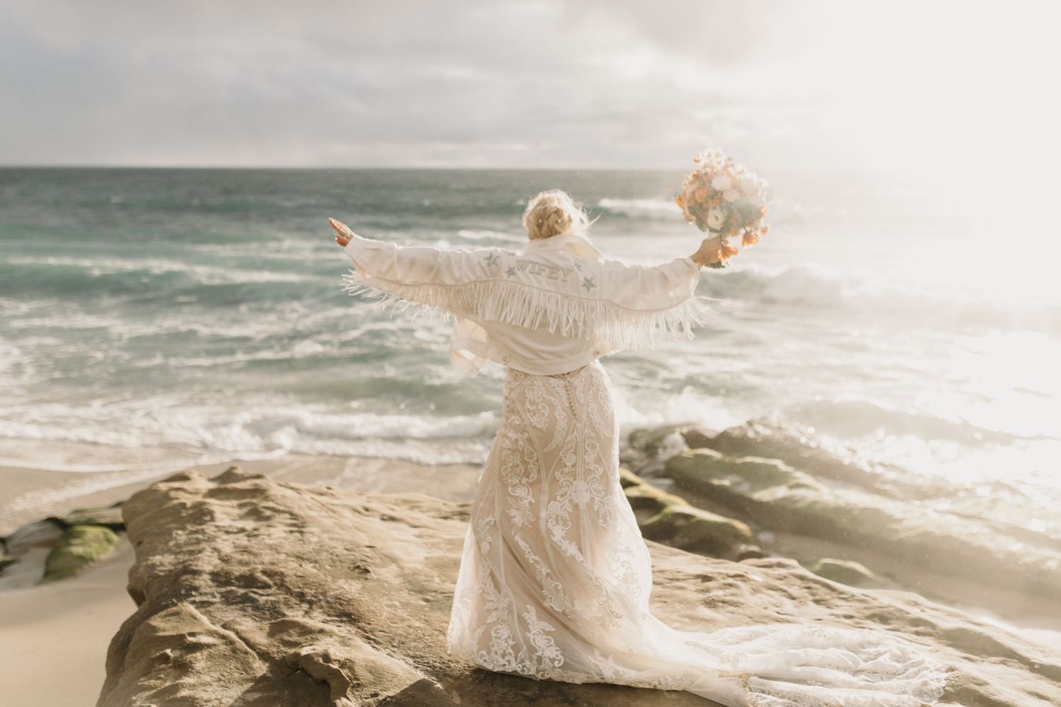 San Diego coast elopement. Windensea beach, La Jolla. Joyful bride in lace gown stands on rocky cliff with arms outstretched facing ocean.