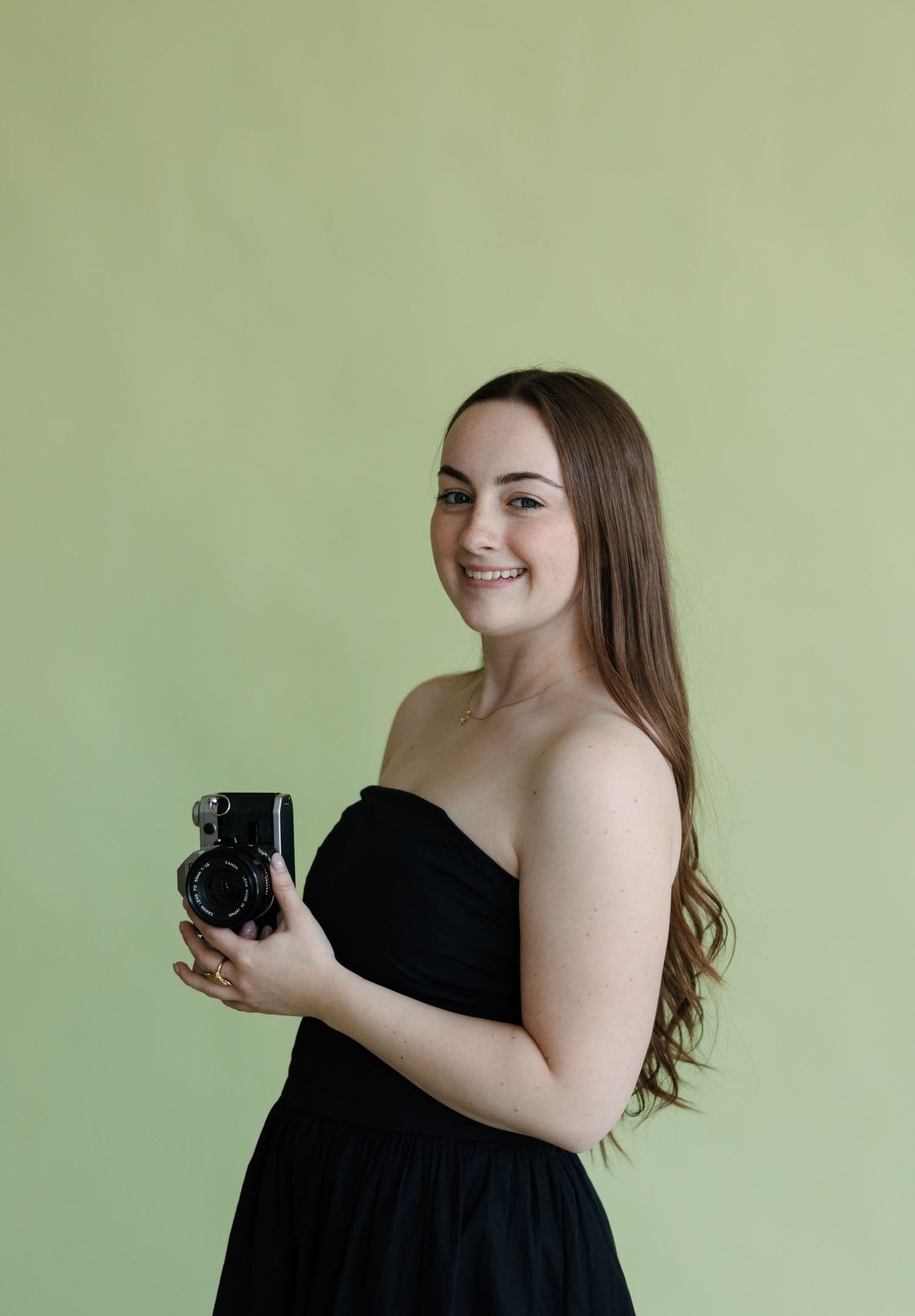 Smiling young woman in black strapless dress holding a vintage camera against a light green background.