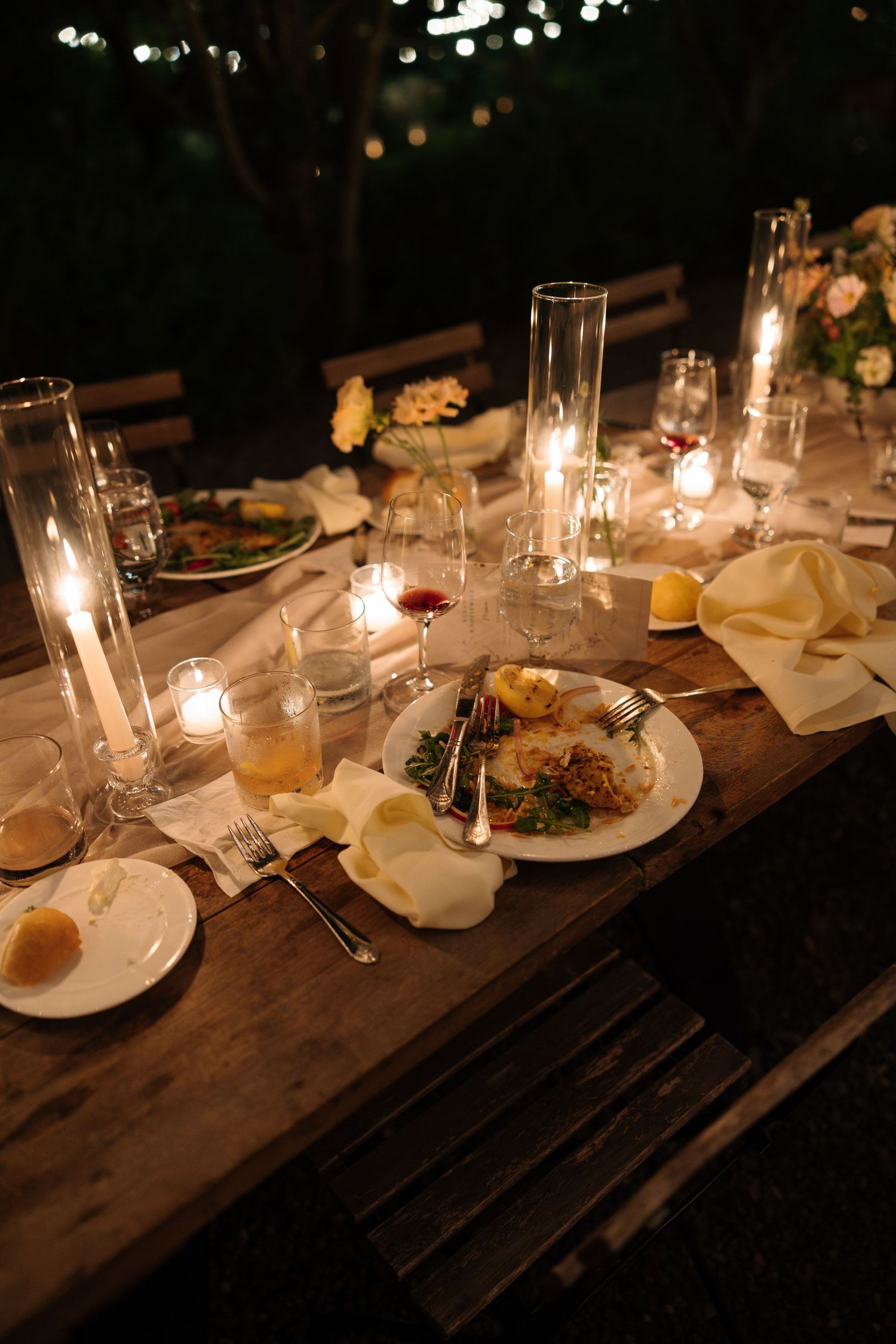 Elegant outdoor candlelit dinner table set with plates of food, glassware, and floral arrangements at night.