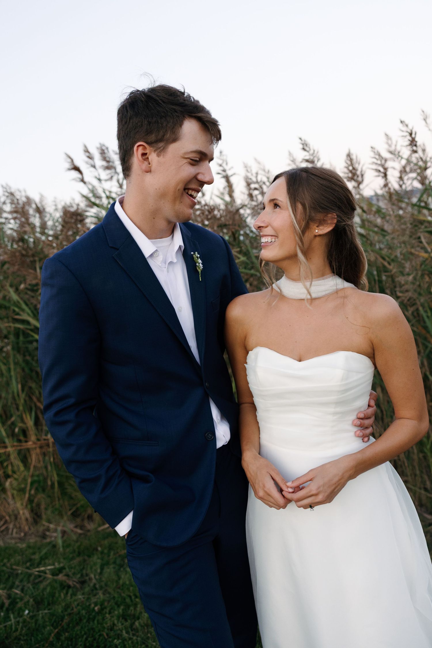 A romantic wedding couple embraces outdoors against a backdrop of tall grasses at sunset.