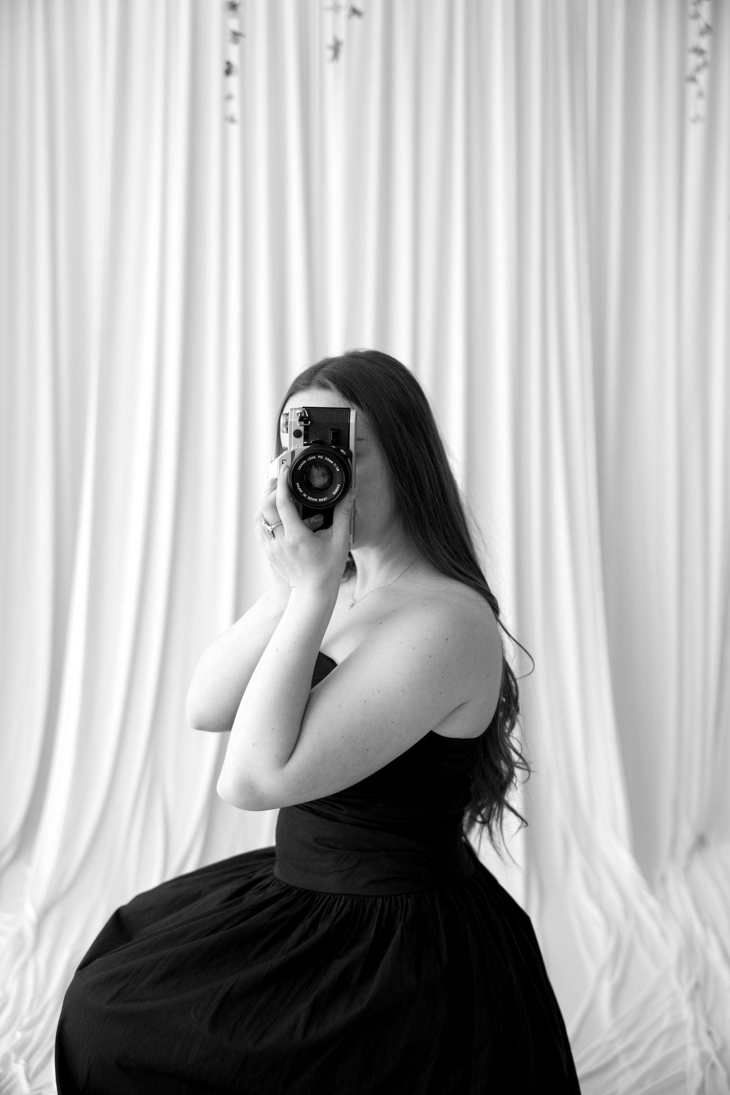 Black and white photo of a woman in a black dress holding a vintage camera up to her face against white curtains.