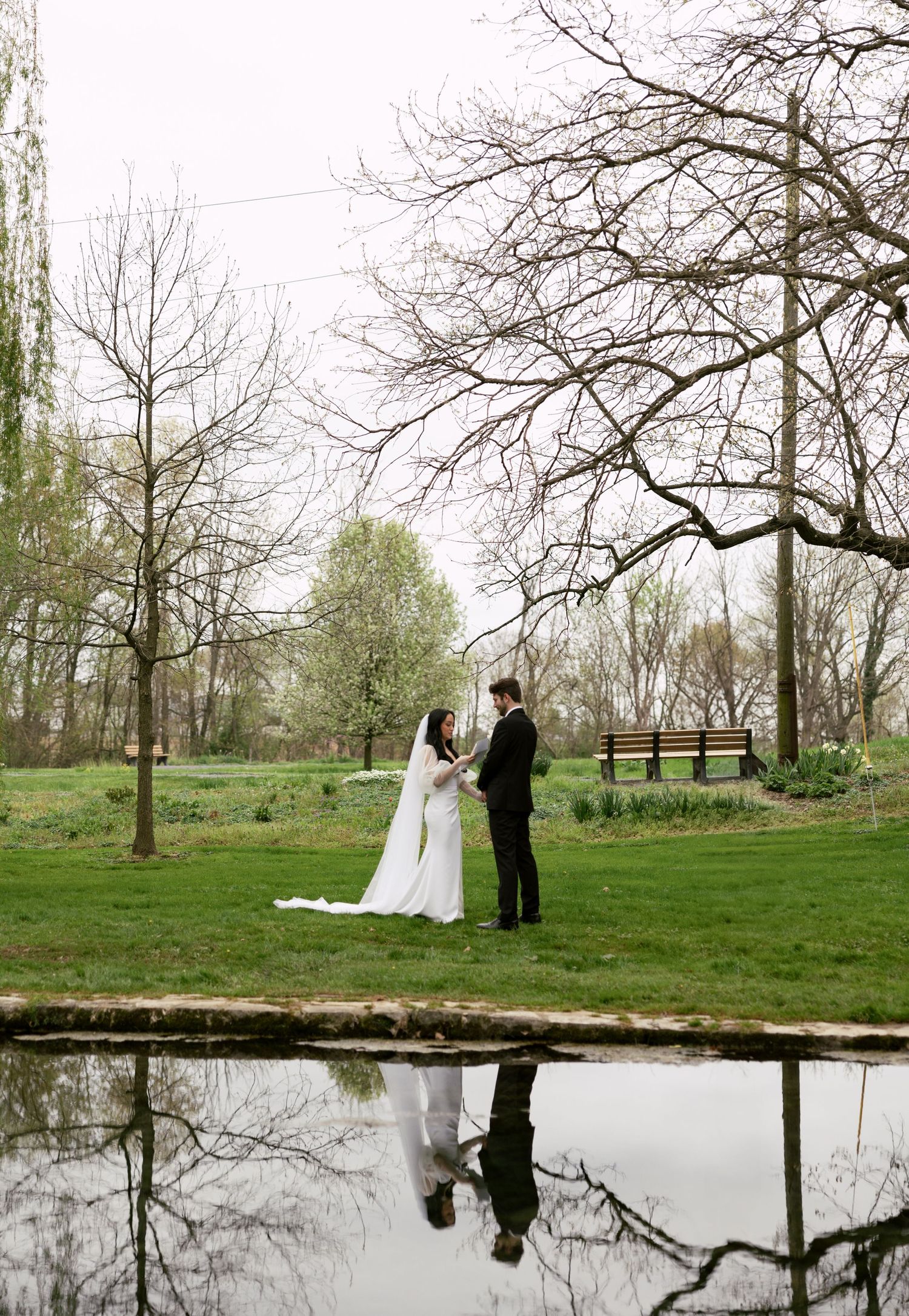 A couple stands reflected in still water beneath bare winter trees.