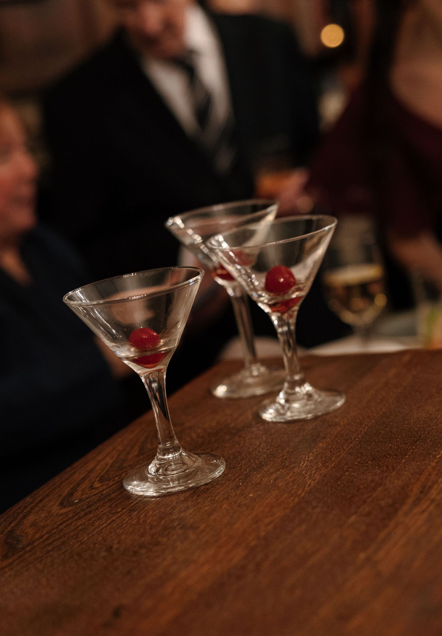 Three classic martini glasses with red cherries sit on a dark wooden bar in a dimly lit setting.