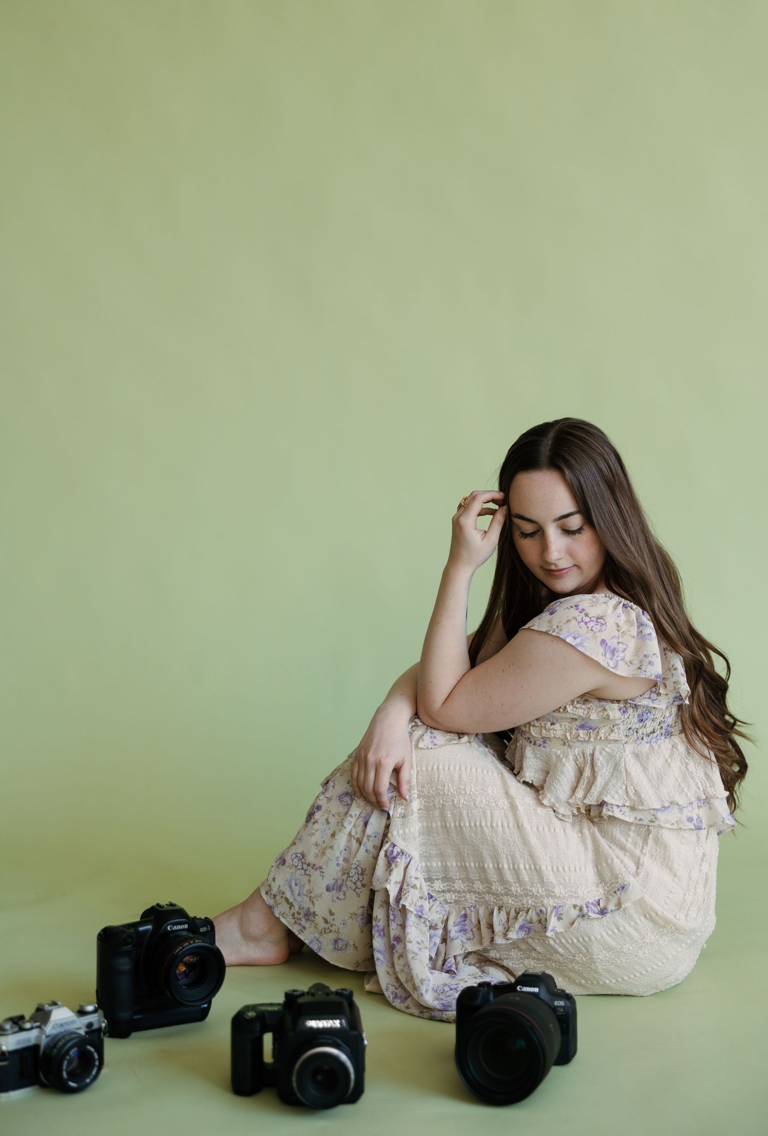 Woman in floral dress sitting on floor surrounded by cameras against green background.