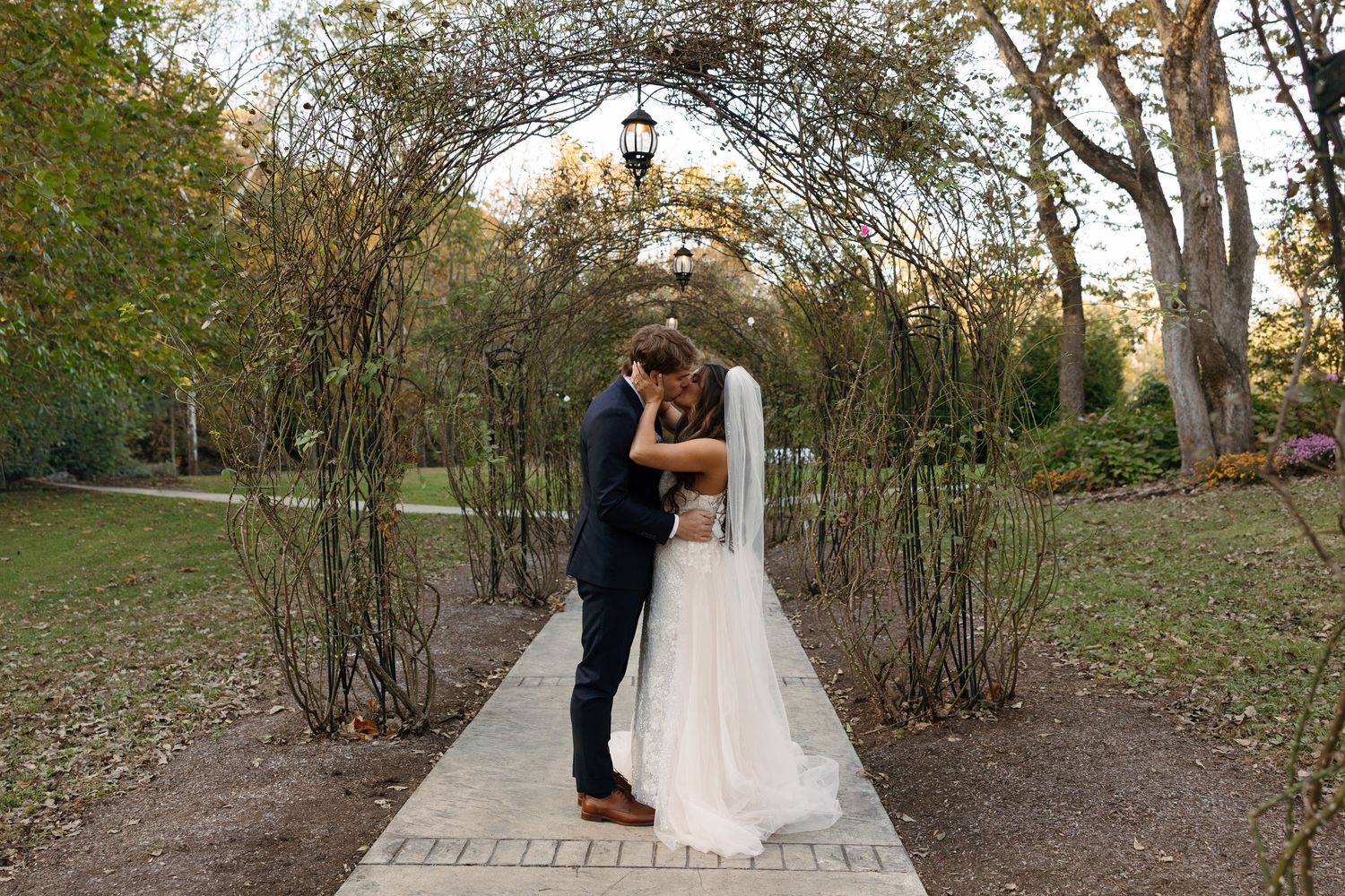 Bride and groom share a romantic kiss under a vine-covered garden arch on a brick pathway surrounded by lush greenery.