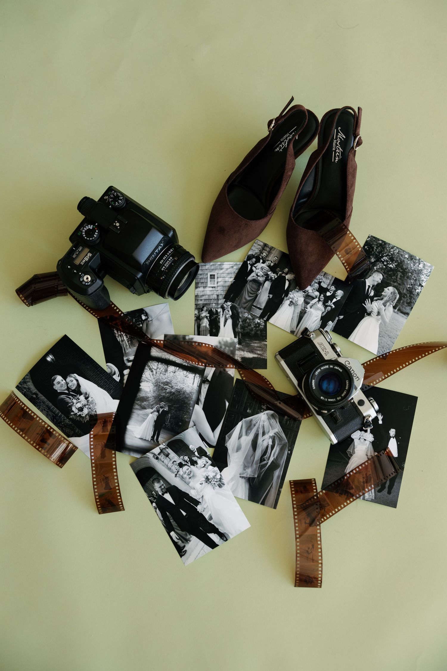 Flat lay of black and white photos, film negatives, two cameras, and brown heels on a light green background.