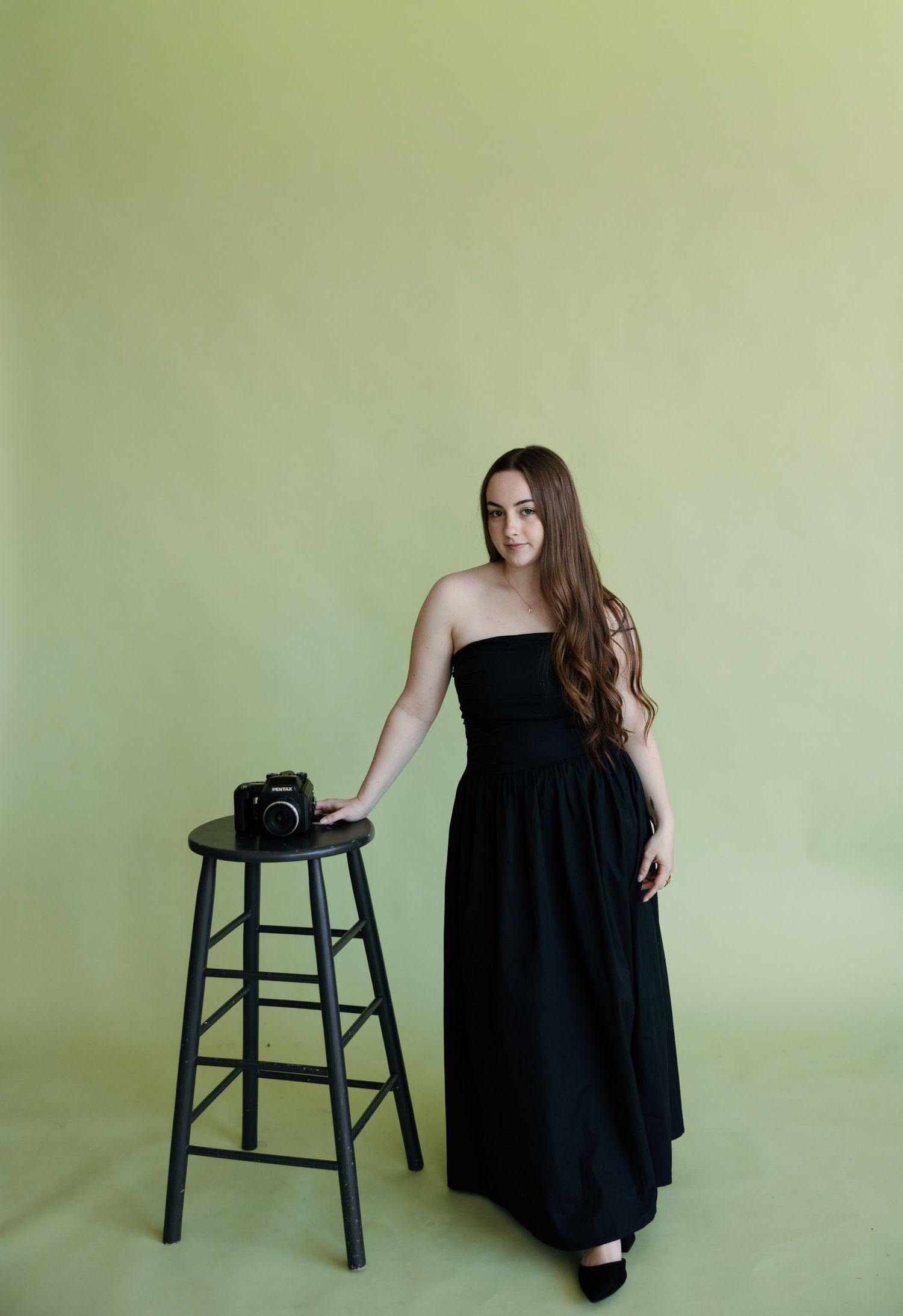 Woman in black strapless maxi dress posing beside a wooden stool with a camera against a green backdrop.