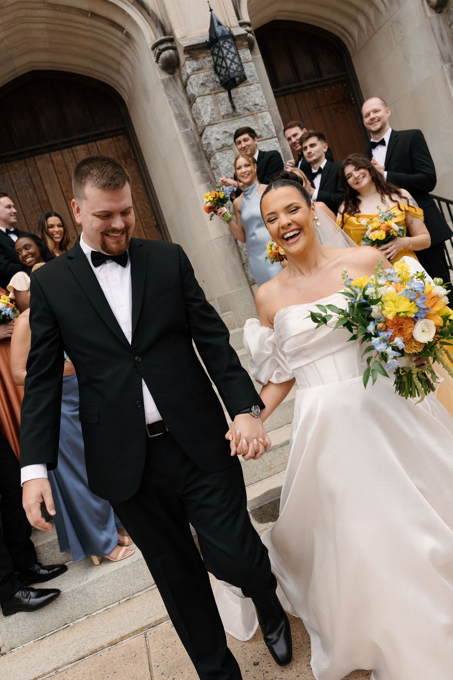Joyful wedding couple exits church with bright yellow sunflower bouquet and formal attire as guests look on.