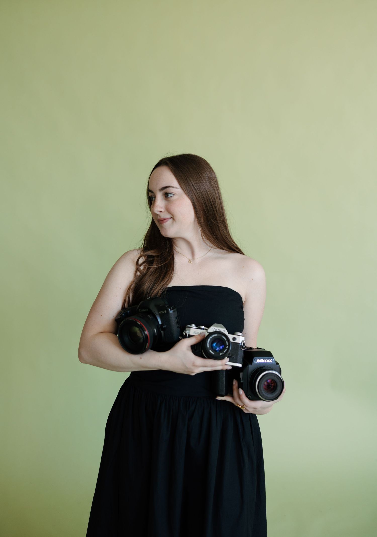 Young woman in black strapless dress holding two cameras, smiling against a light green background.