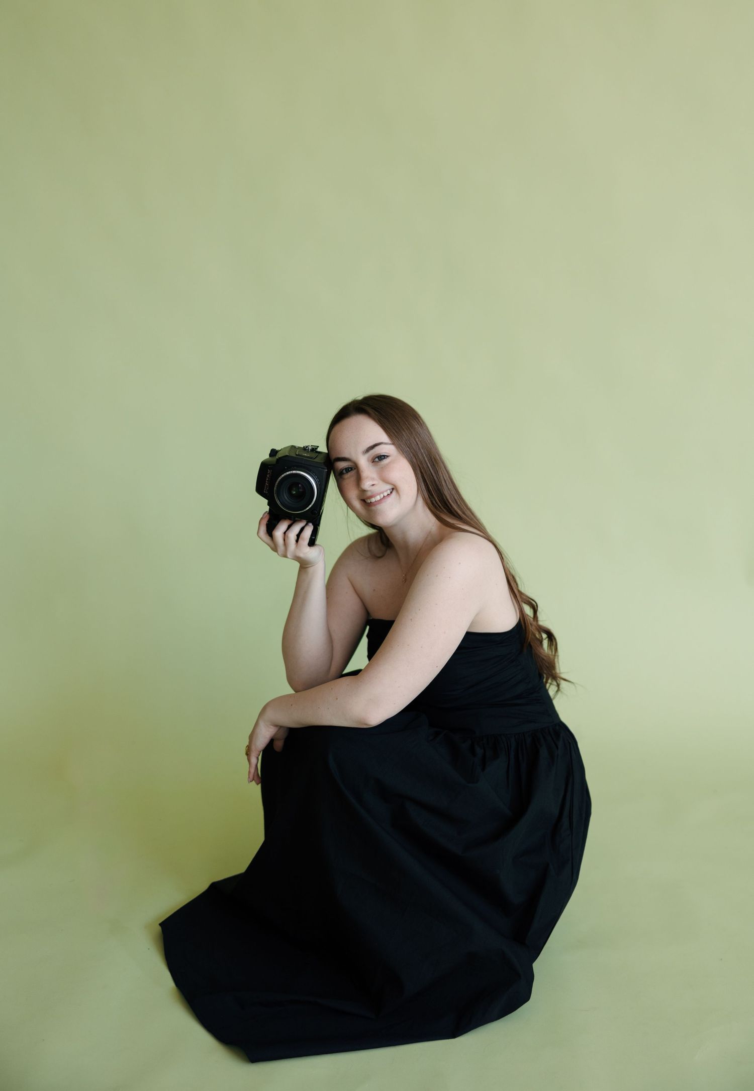 Young woman in black dress sitting on floor holding camera, smiling against light green studio backdrop.