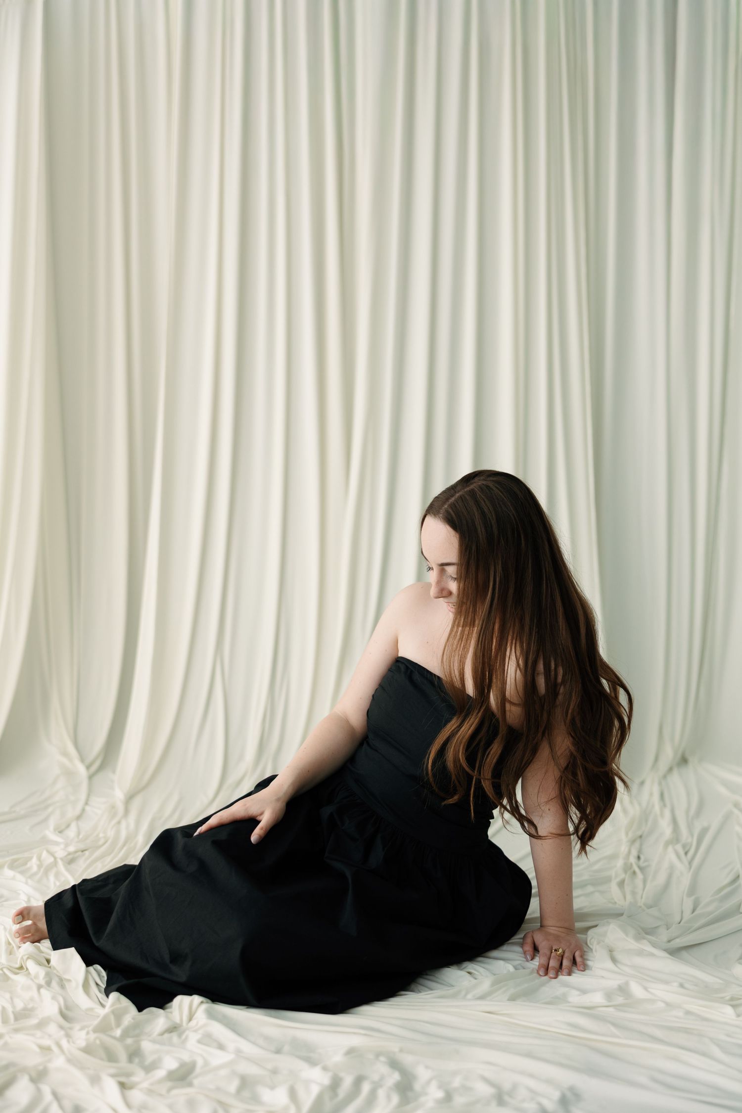 Young woman with long brown hair in black strapless dress sitting gracefully on white fabric backdrop.