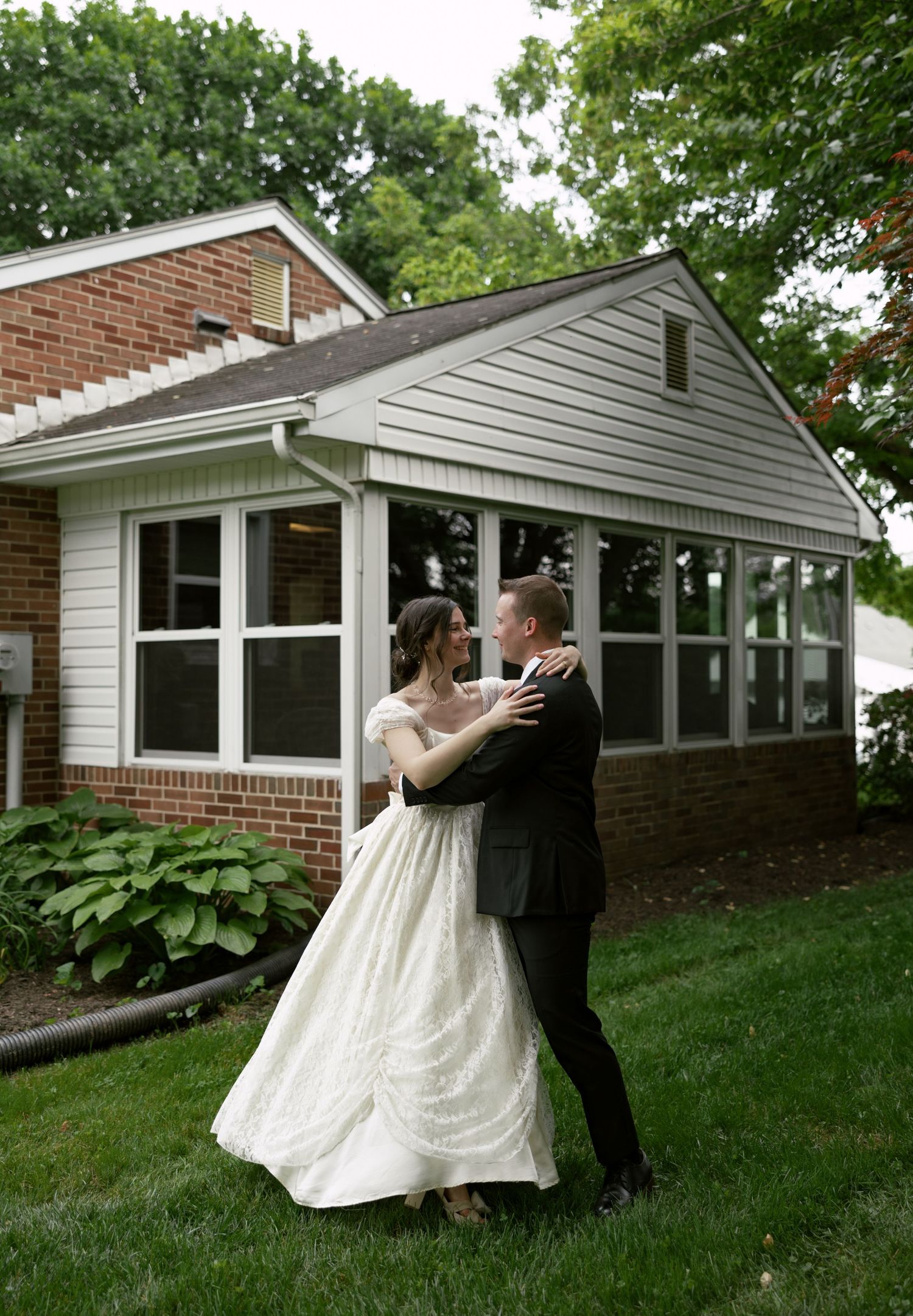 A couple dances on the lawn in front of a brick and white siding ranch-style house.