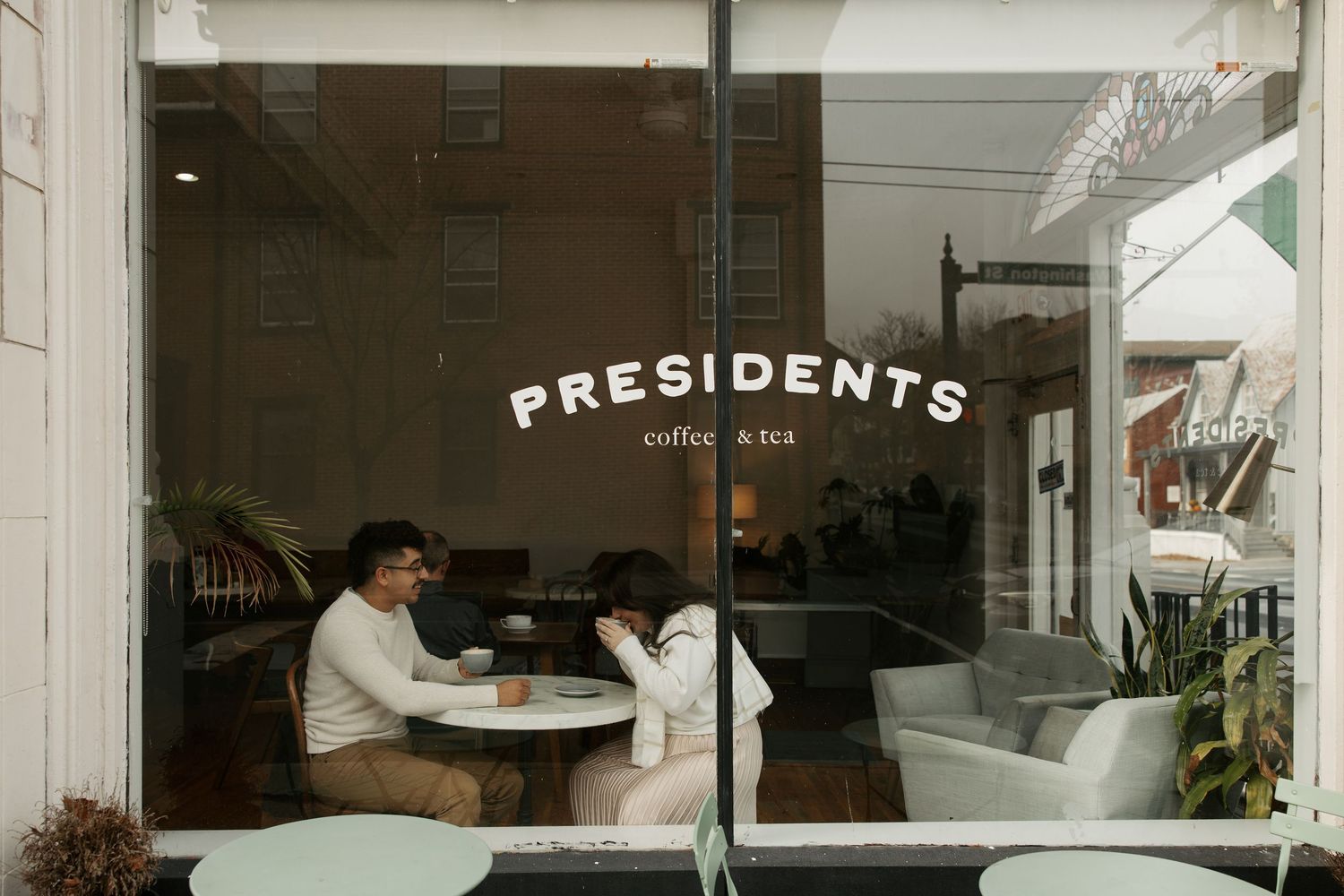 Two people sit at a table inside a coffee shop window with 'Presidents' text signage visible from outside.