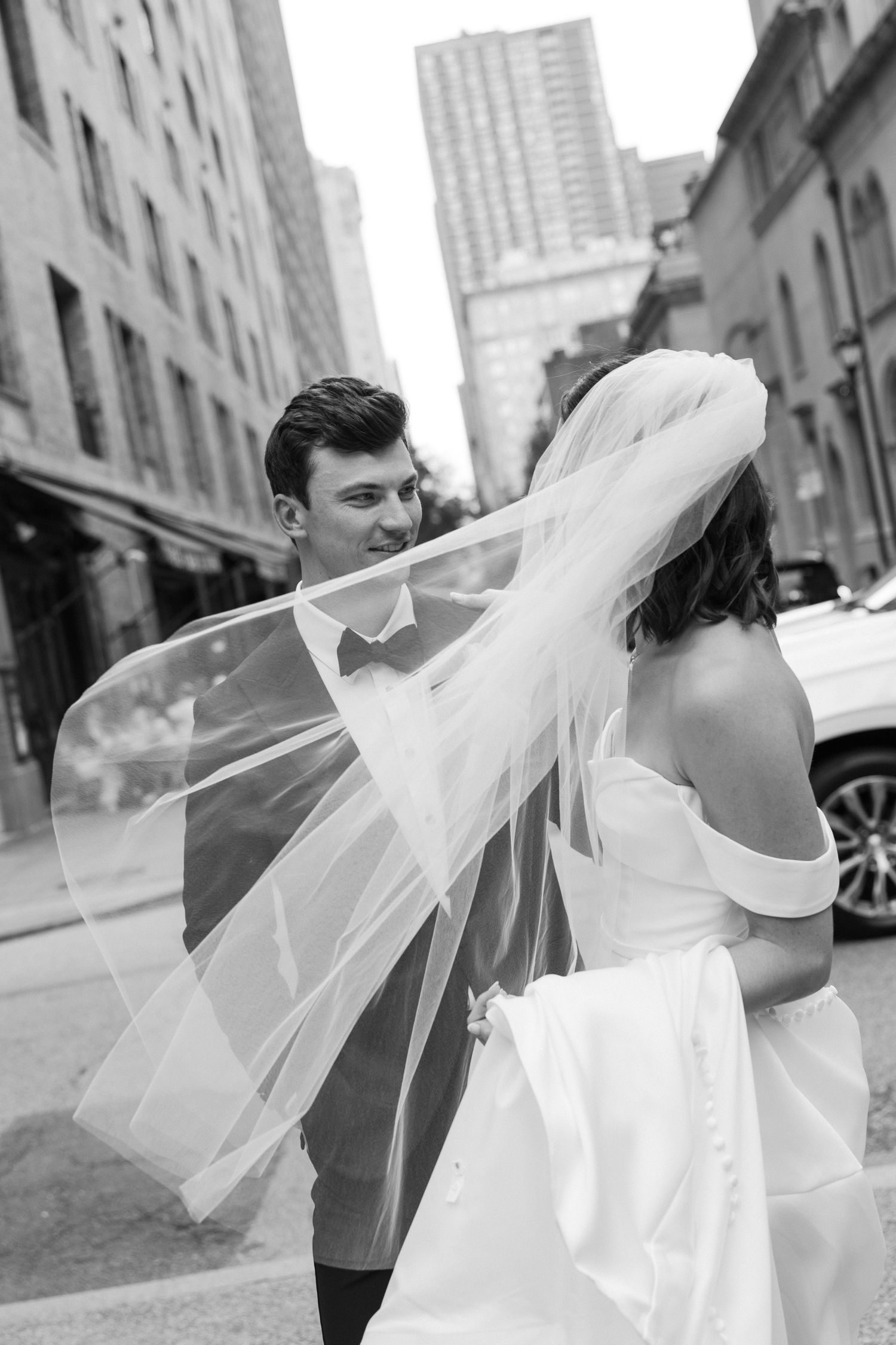 Black and white photo of a bride and groom walking city streets, her veil flowing in the wind.