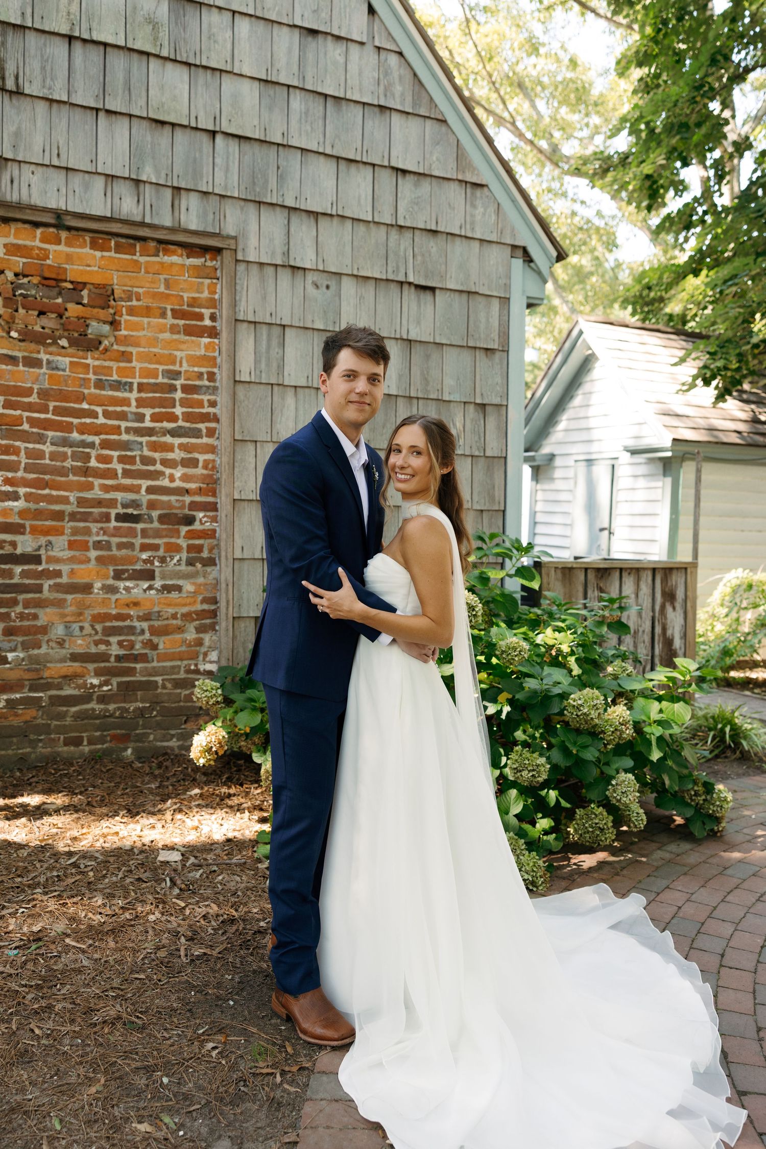 A wedding couple shares a romantic moment outside a rustic barn with brick and wood siding on a sunny day.