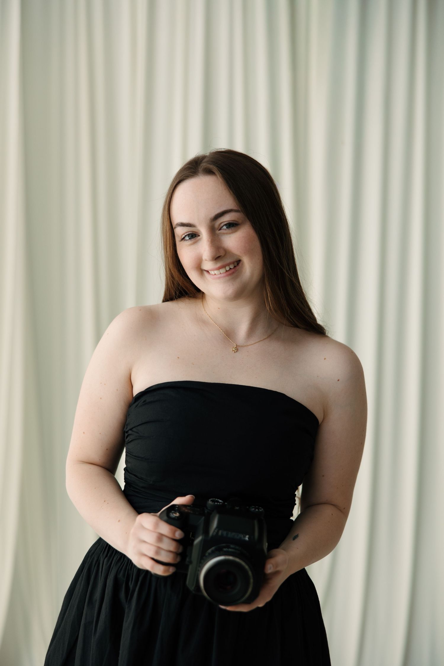Smiling young woman in black strapless dress holding a DSLR camera against a white curtain backdrop.