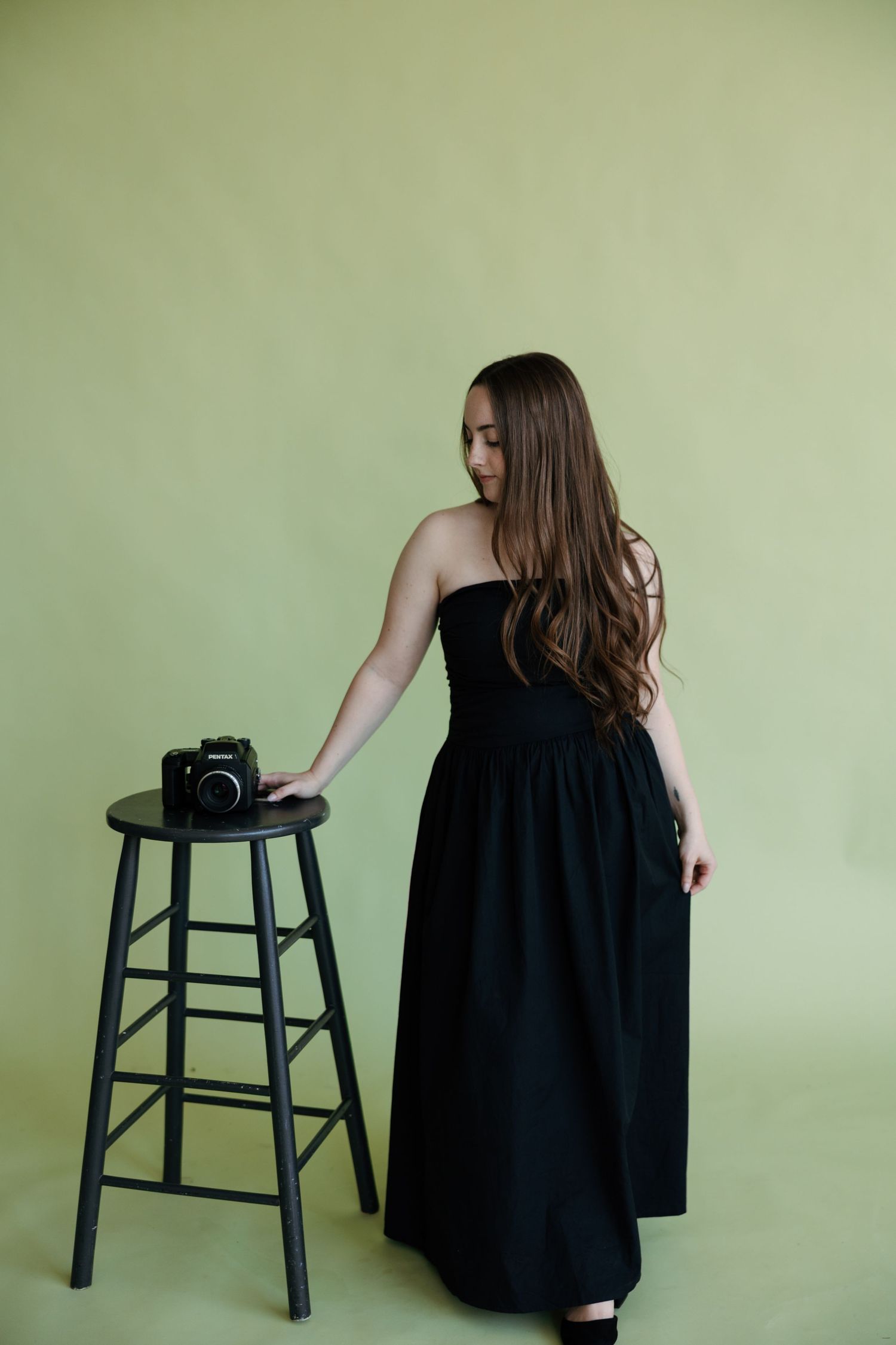 Young woman in black strapless maxi dress stands beside a wooden stool with a vintage camera on green background.