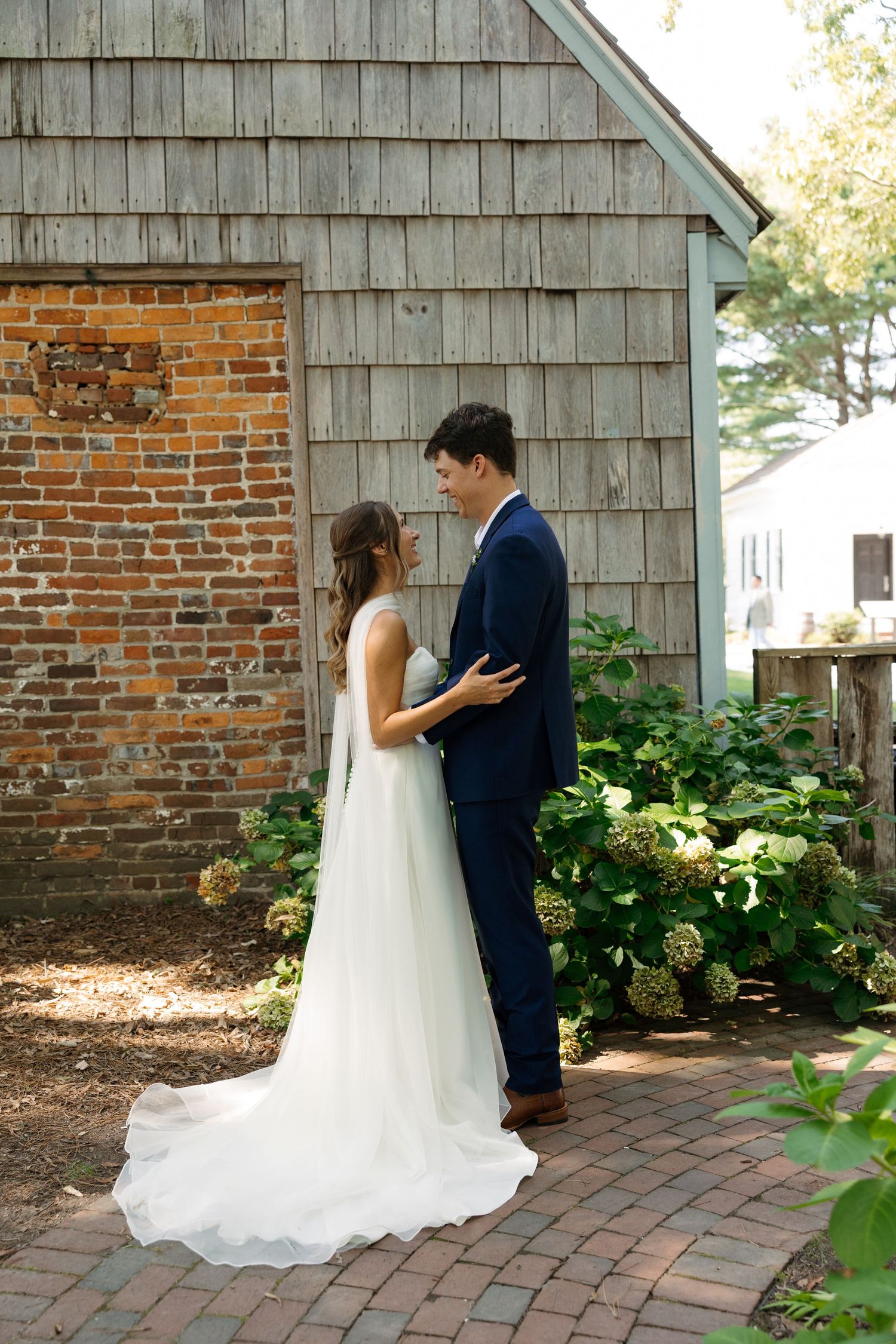 Bride and groom share a tender moment outdoors near a rustic brick and wood building surrounded by lush hydrangeas.