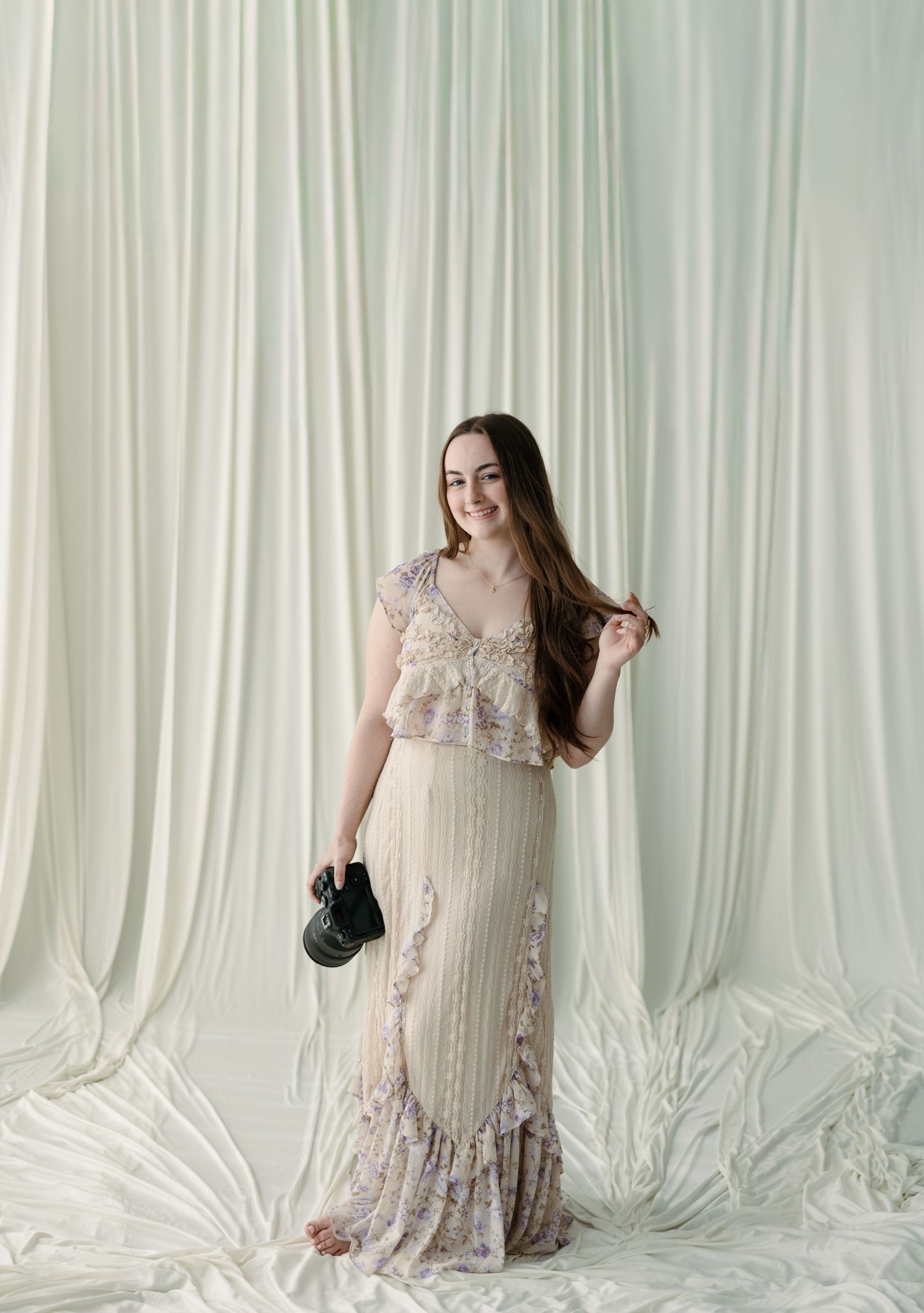Young woman smiling in a floral maxi dress, holding a black bag, posing against white draped fabric backdrop.