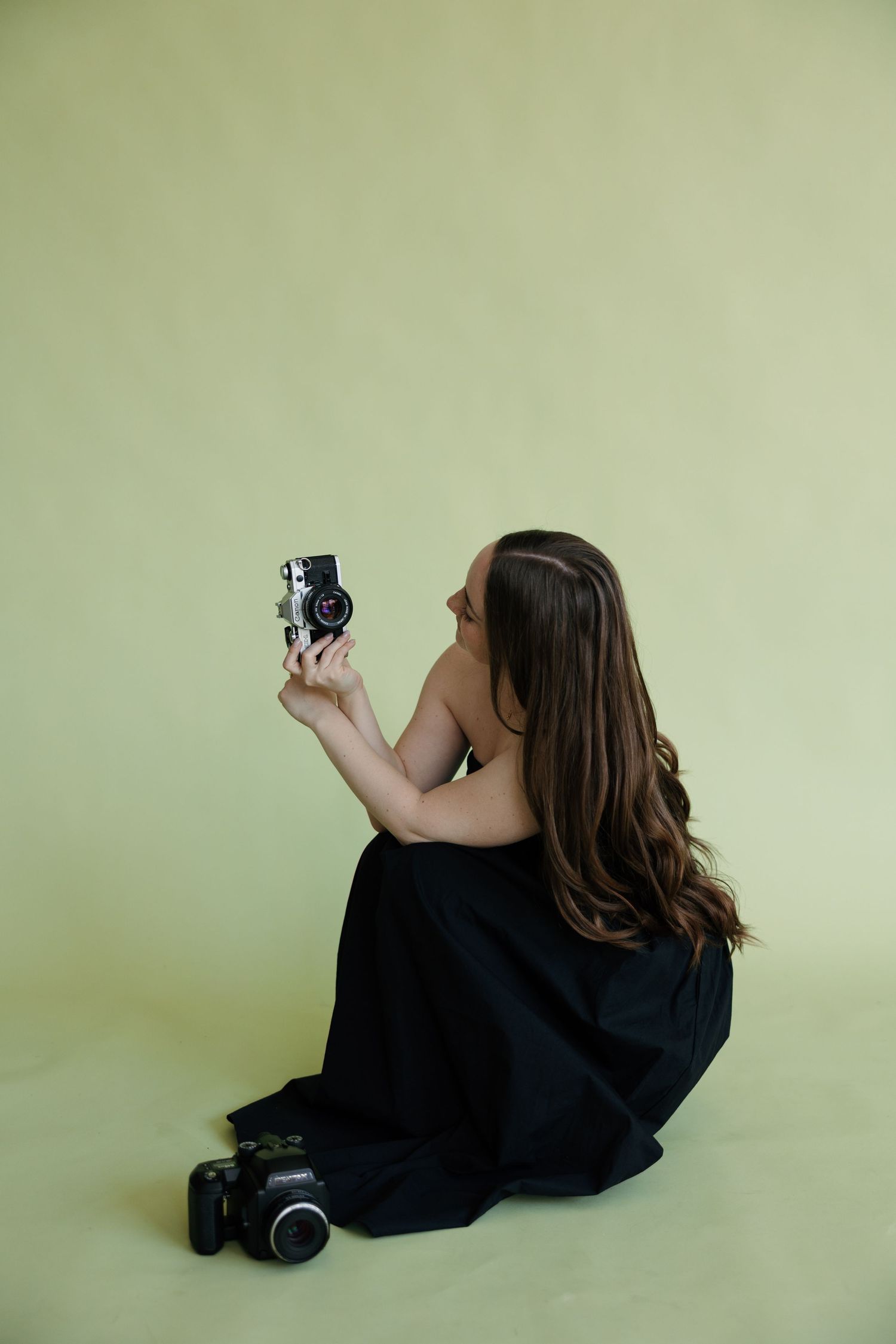 Woman in black dress sitting on floor holding vintage camera up, with DSLR camera beside her on light green background.