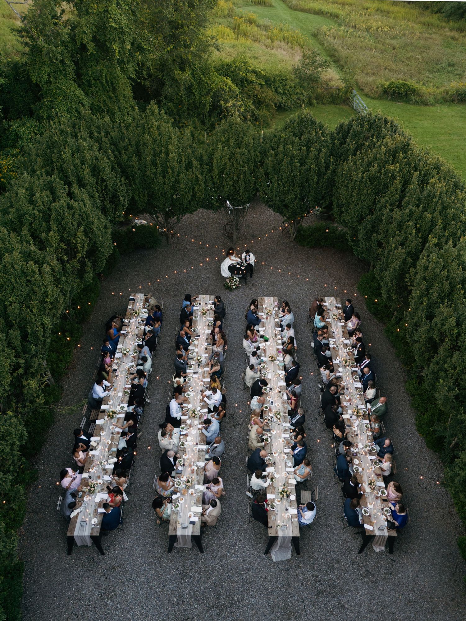 Aerial view of an elegant outdoor wedding reception with long banquet tables arranged symmetrically among lush green hedges at dusk.