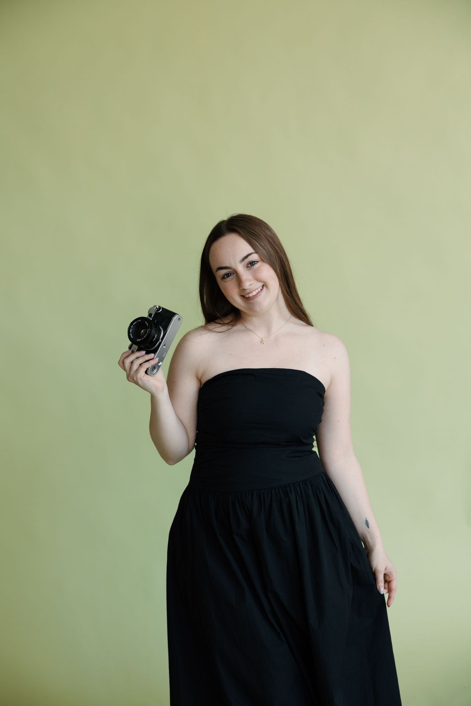 Smiling young woman in black strapless dress holding a vintage camera against a light green background.