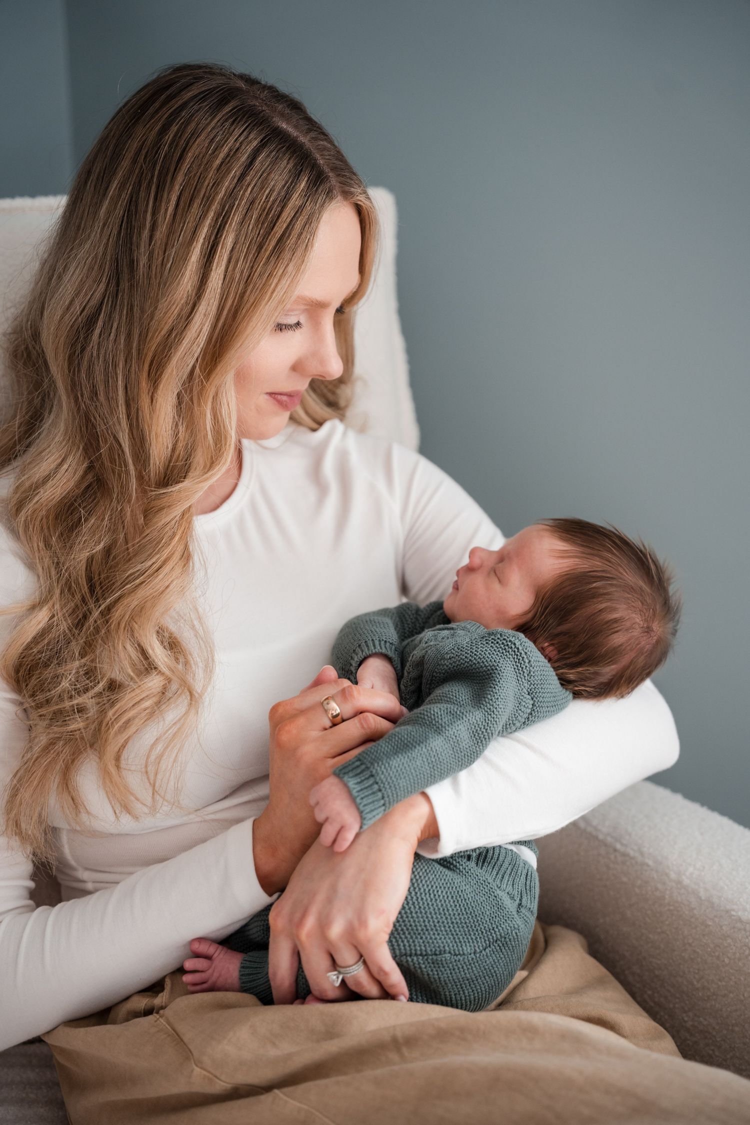 A young mother with long blonde hair holds her sleeping newborn baby dressed in a green knit outfit while sitting in a chair.