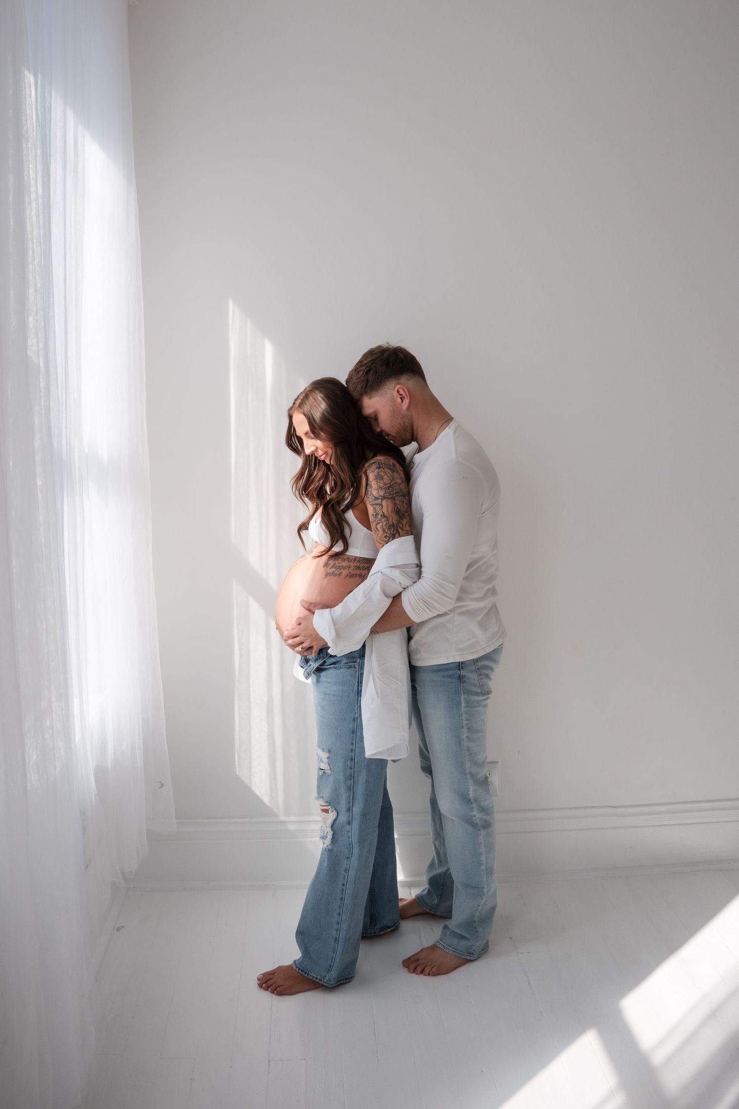 Couple embracing in white room, pregnant woman in ripped jeans and white top, man hugging her from behind near window.