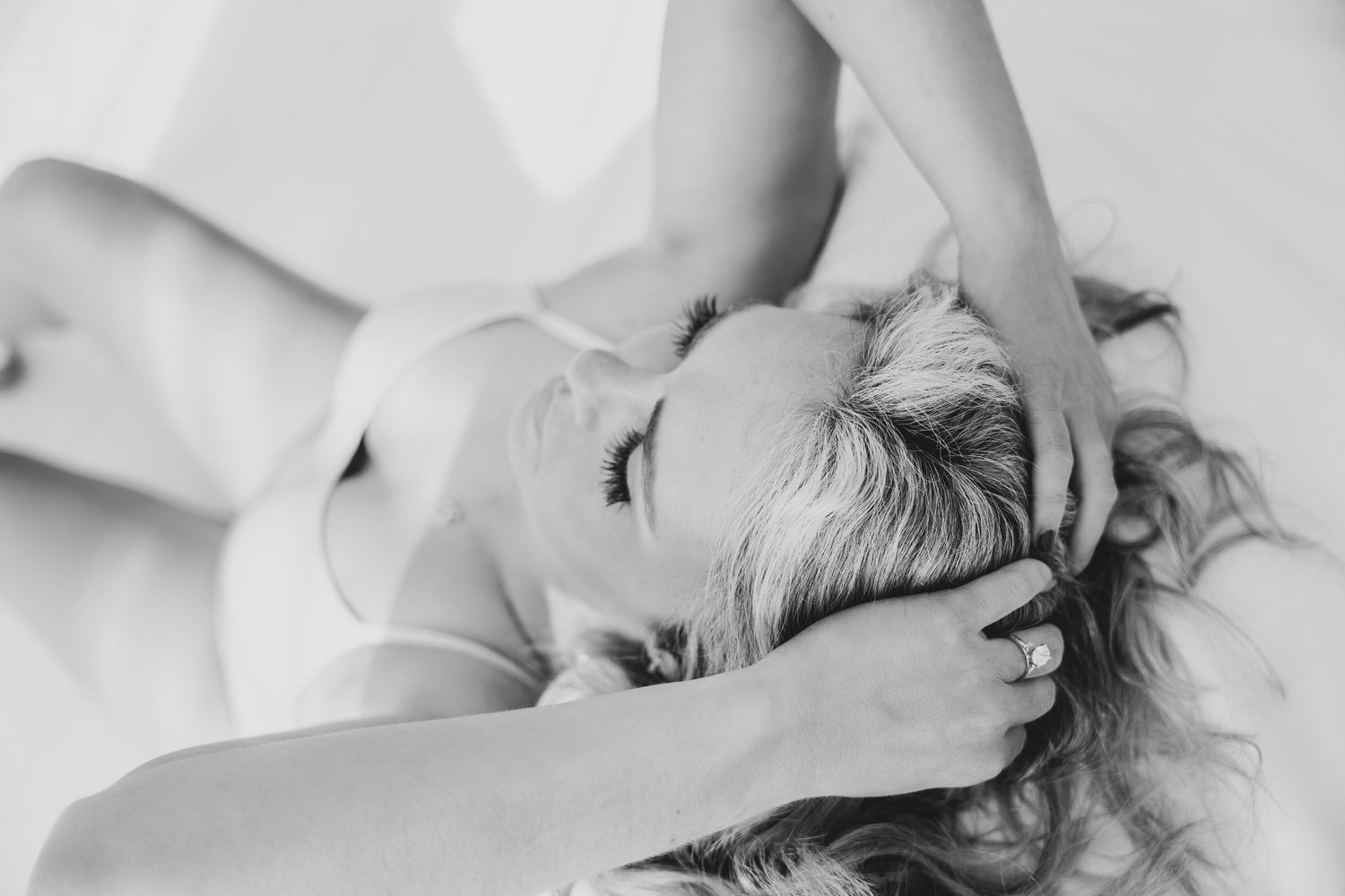 Black and white boudoir photo of a woman lying down with her hands in her hair, wearing lingerie.