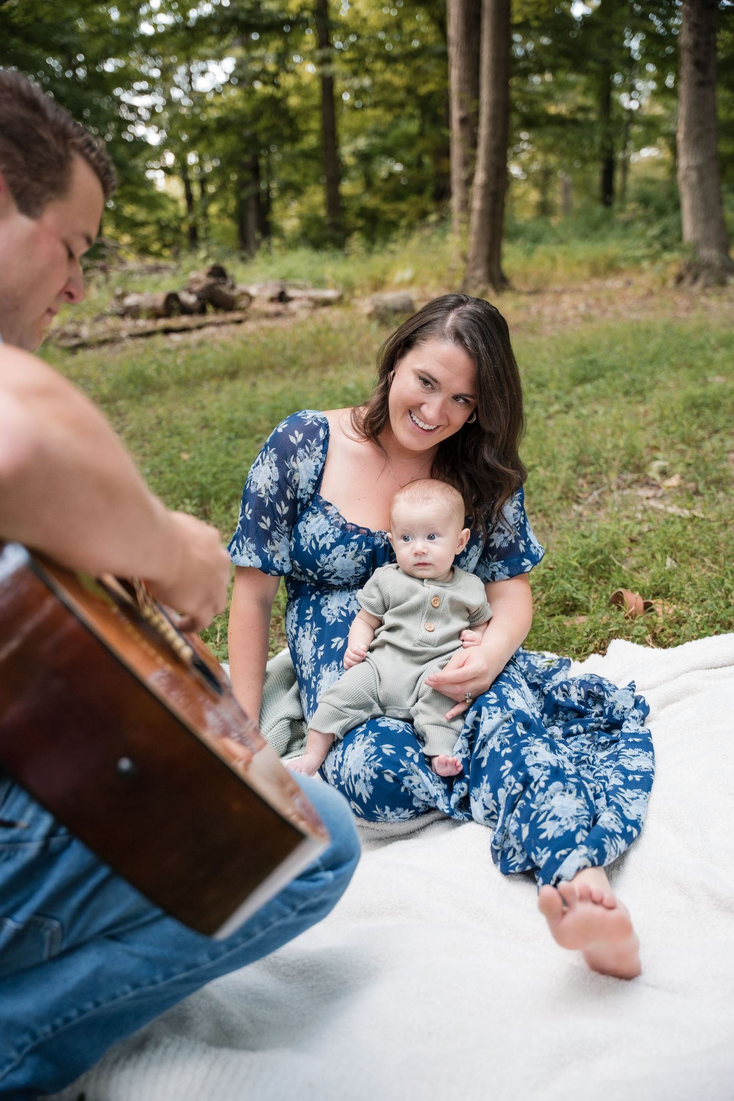 A smiling mother holds her baby on a blanket outdoors while a man plays guitar in a lush green park.