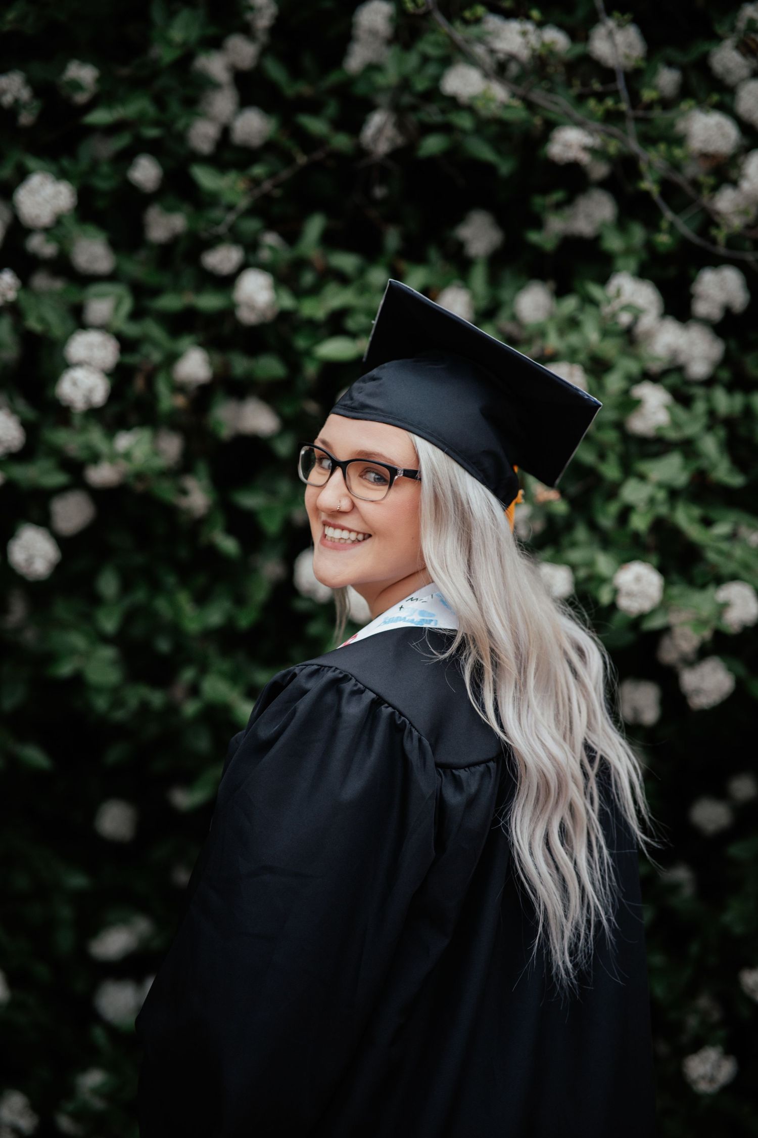 Graduate in black cap and gown with blonde hair stands in front of white flowering bushes and smiles over shoulder.