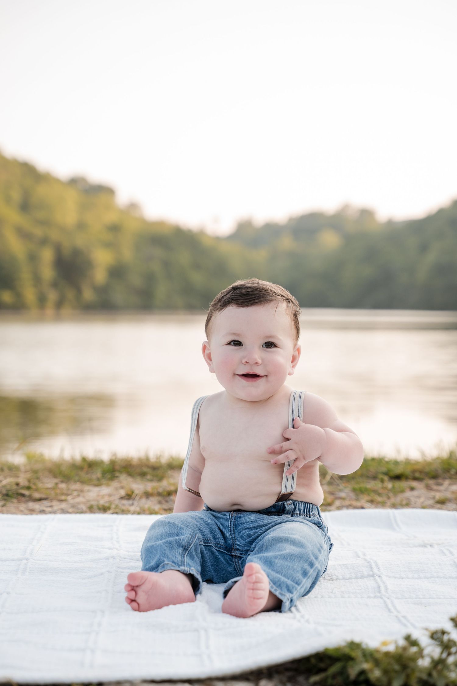Smiling baby boy in jeans and suspenders sitting on a white blanket by a serene lake outdoors.