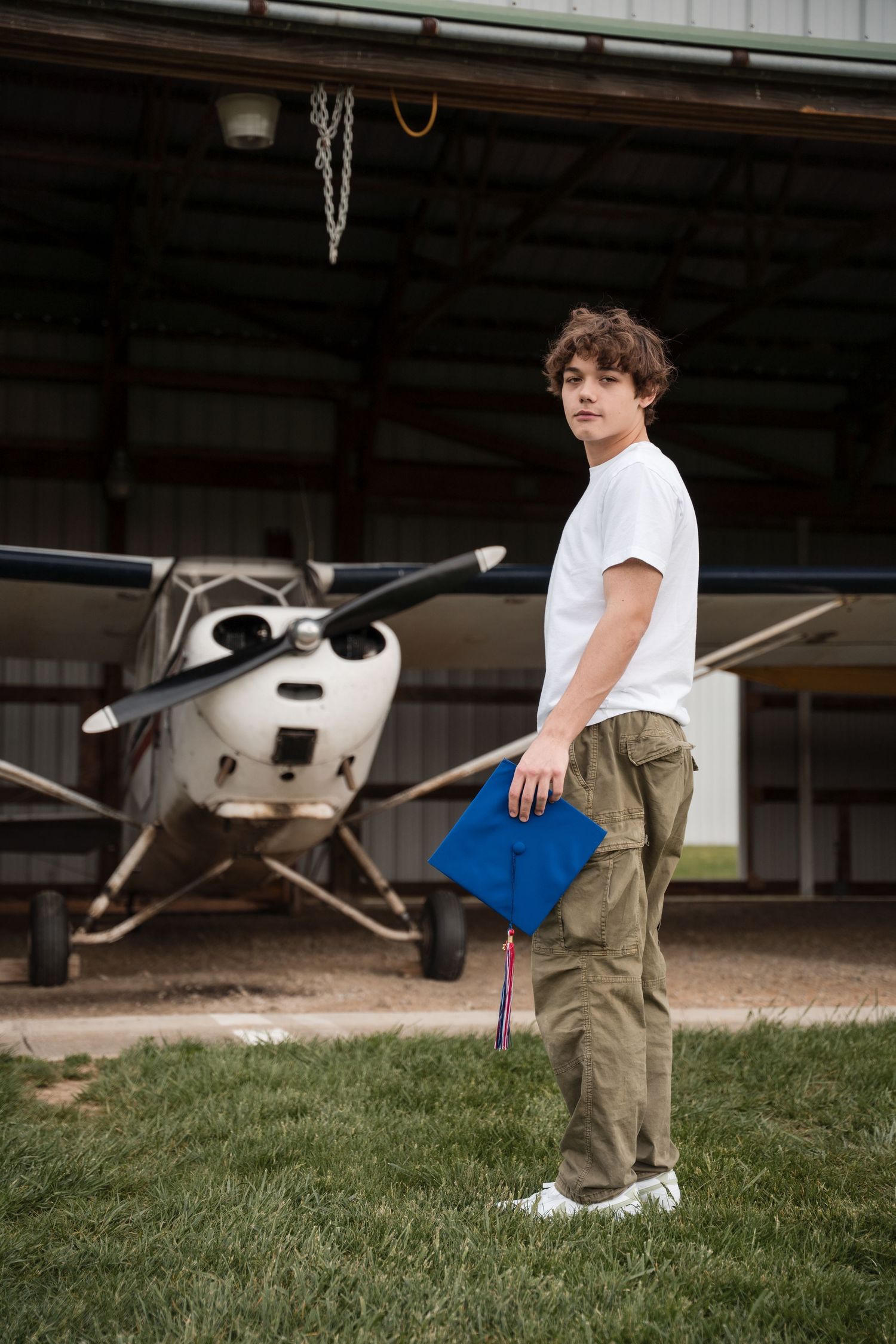 Young man in white t-shirt and cargo pants holds blue graduation cap in front of small aircraft in hangar.