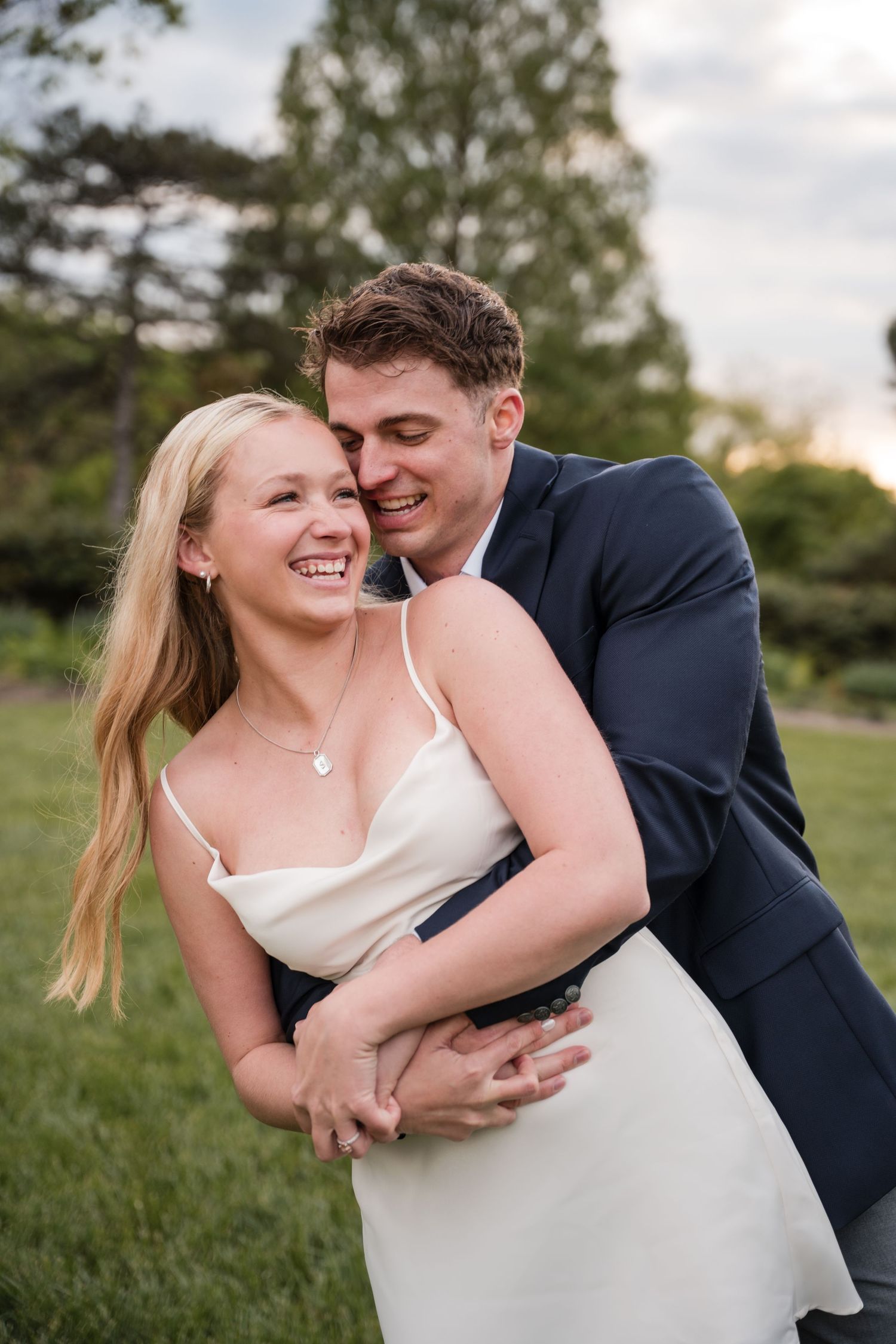 A smiling blonde bride in a white dress embraced by a groom in a navy suit outdoors at sunset.