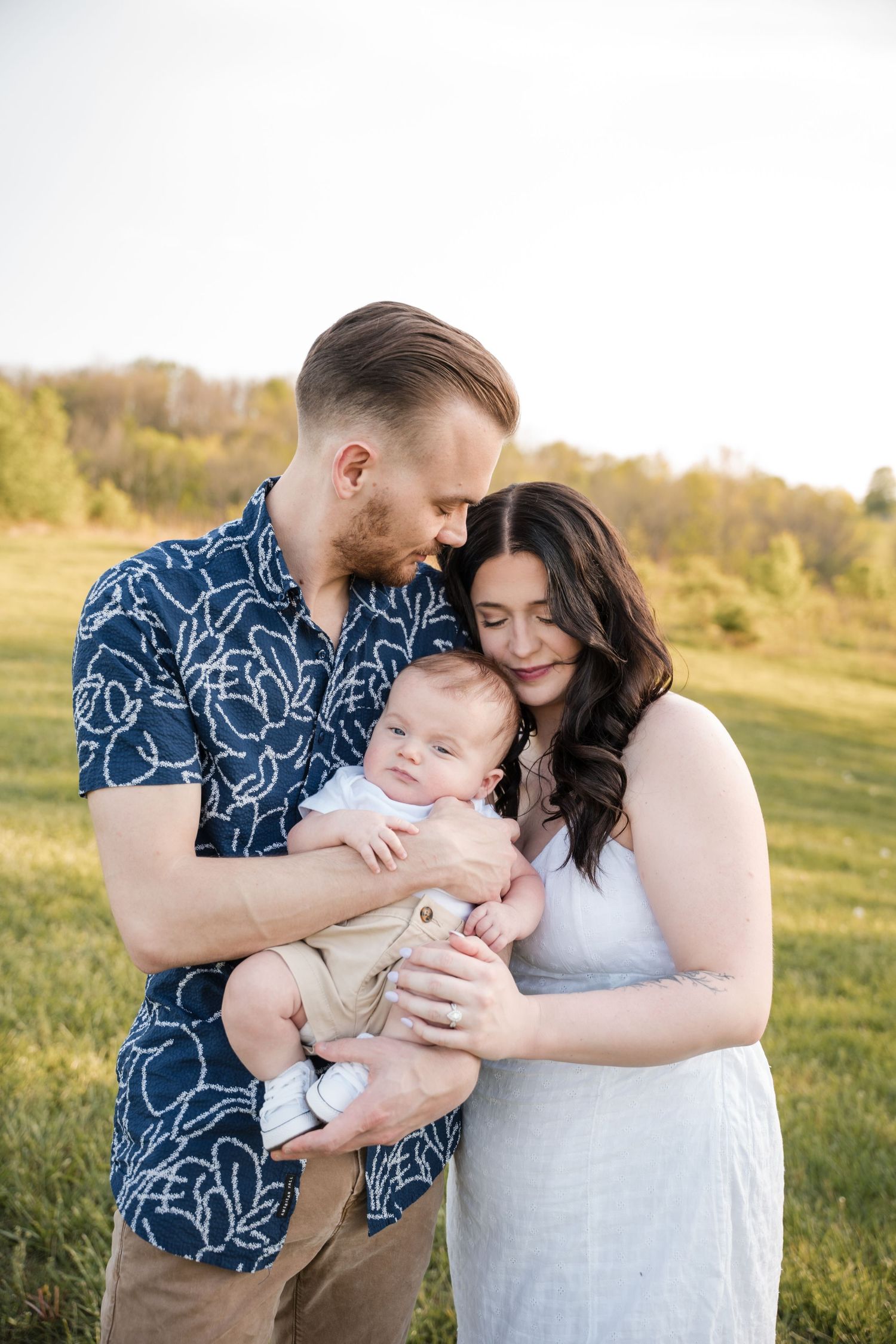 Couple in casual attire tenderly holds their infant in a sunlit meadow at golden hour.