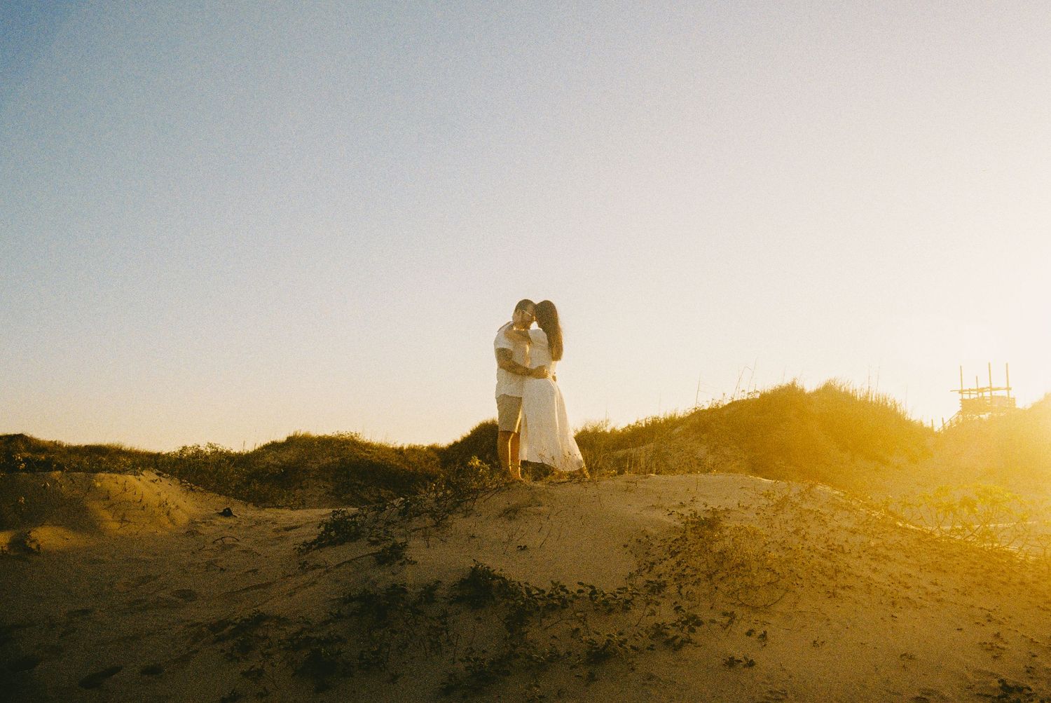 Couple embracing at golden hour on sandy beach dunes, bathed in warm sunset light.