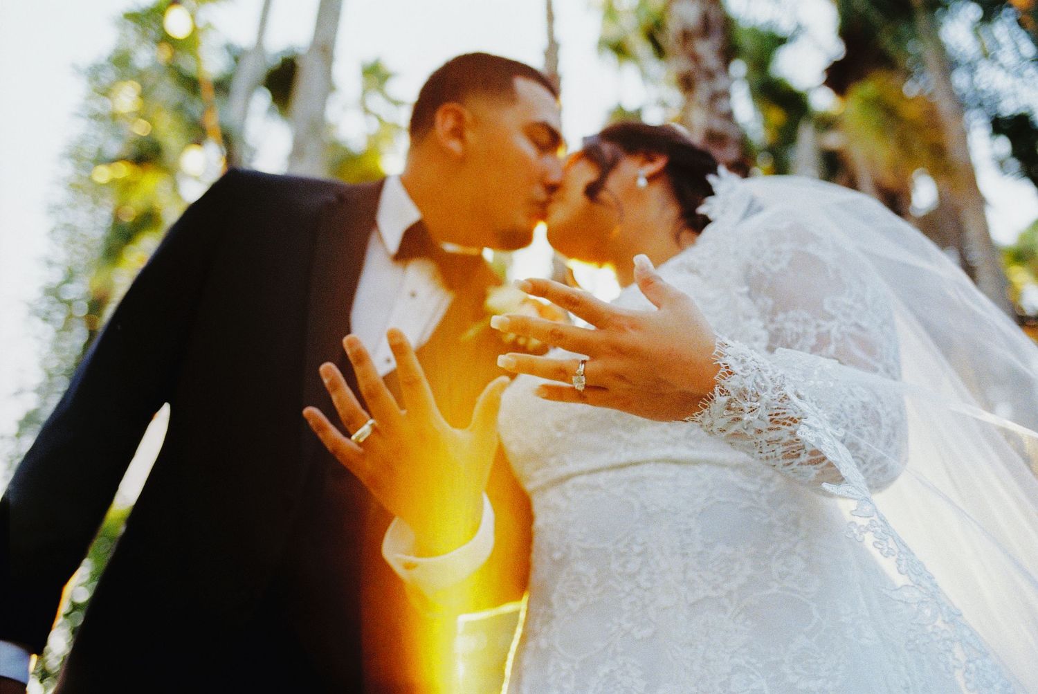 Newlywed couple sharing a kiss while displaying wedding rings, with lush green trees in the background.