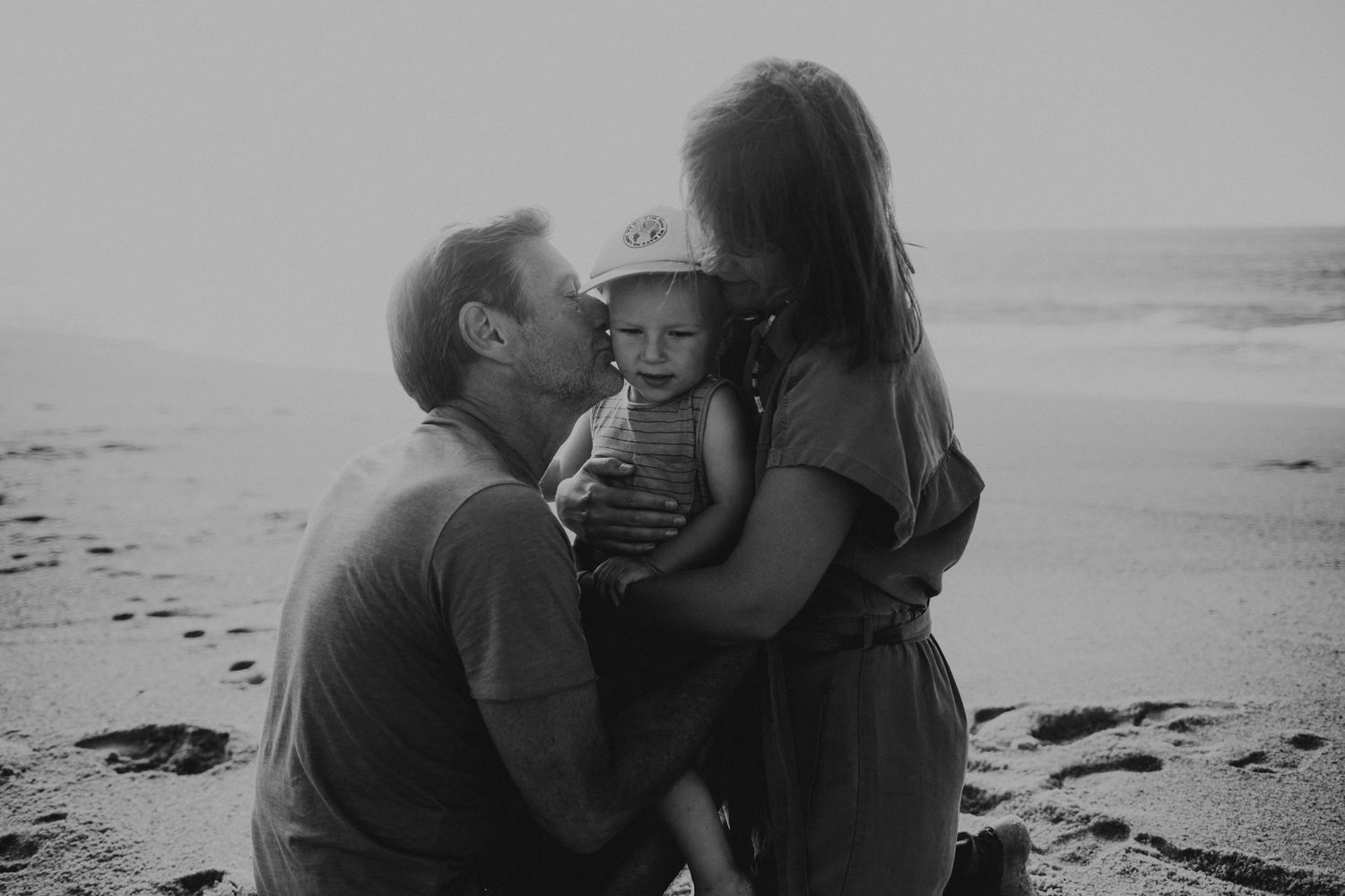 Foto preto branco de casal com o filho no meio a receber um beijo durante sessão fotográfica de familia em Praia Do Salgado Portugal