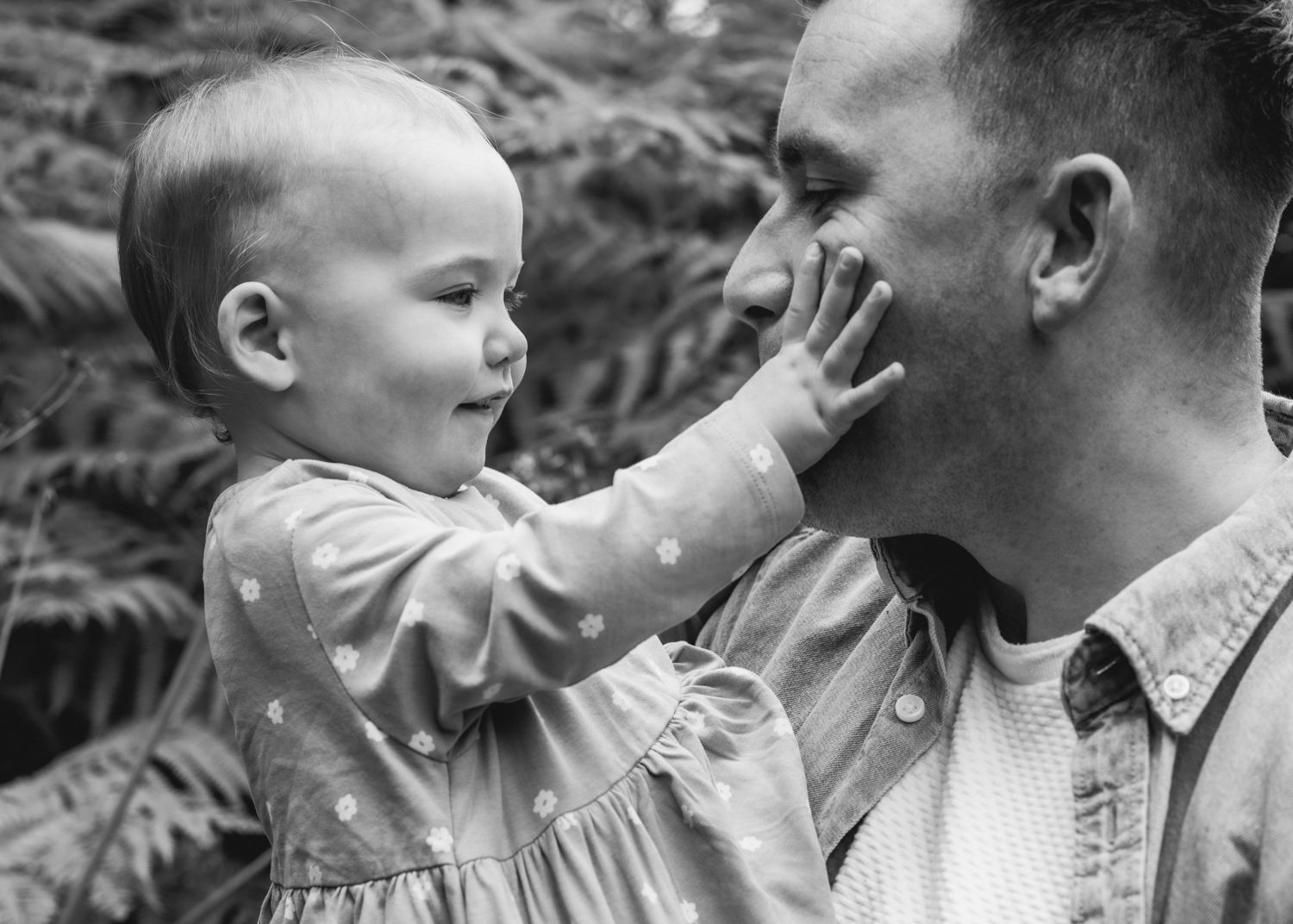 Black and white close-up of a father holding his baby, the child gently touching his face as they share a tender moment outdoors.