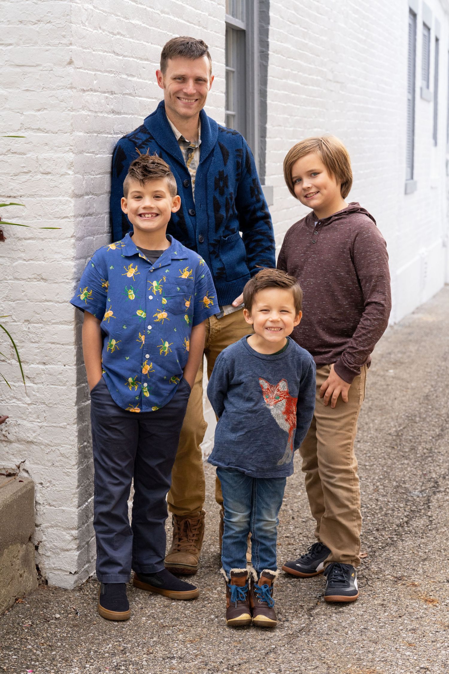 Dad with three sons standing together and smiling against white brick wall in Worthington Ohio, by Jamie Macey Photography