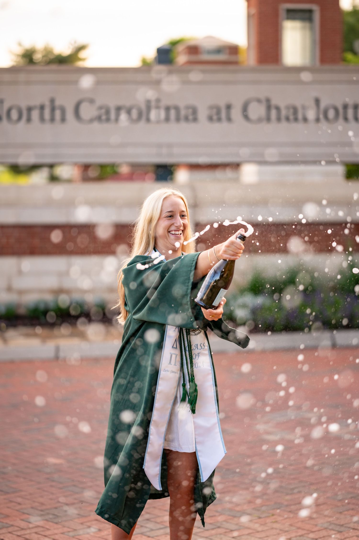 A graduate in a green jacket celebrates with champagne spray on a brick courtyard at UNC Charlotte.