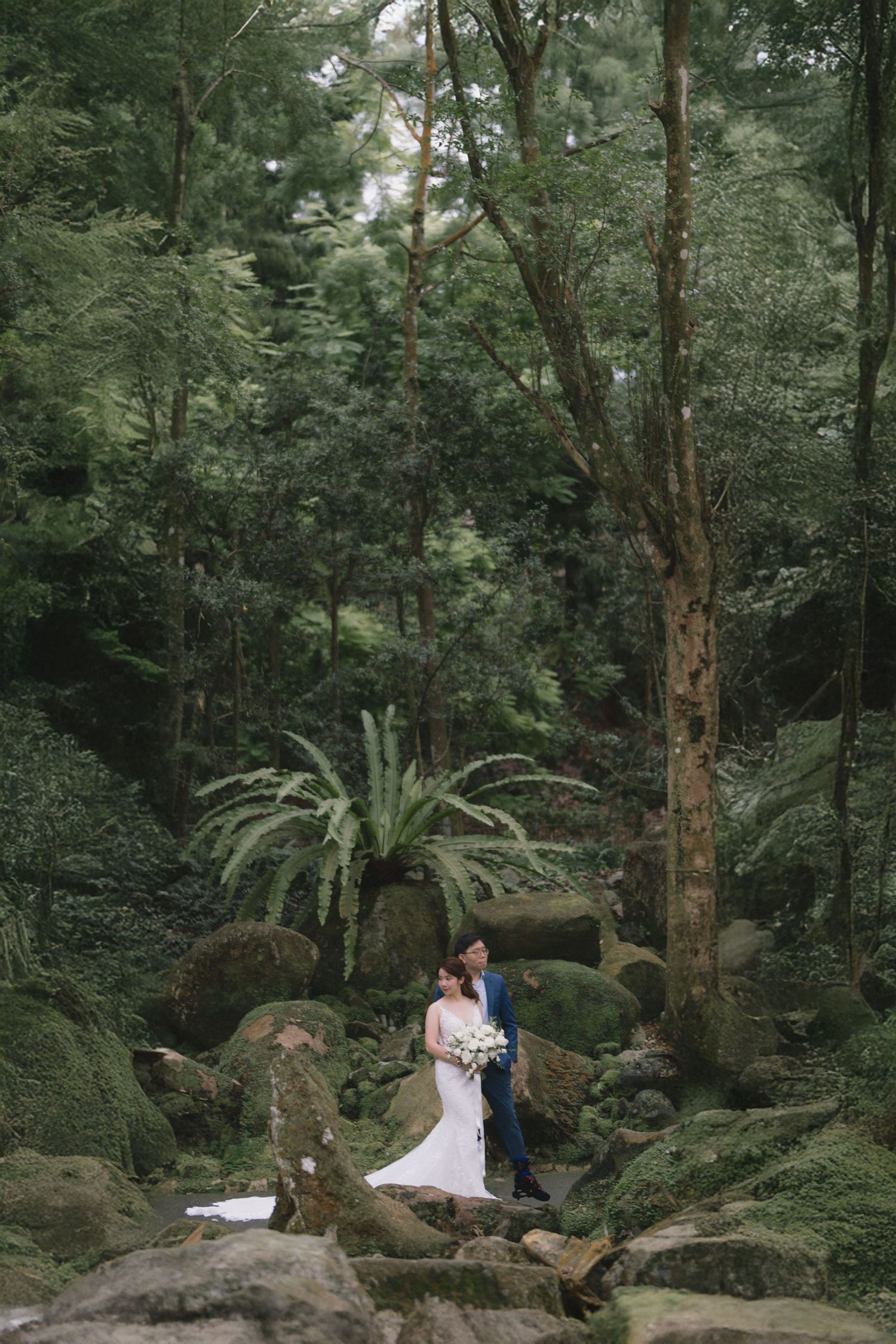 Bride and groom stand among moss-covered rocks in a lush green forest, bride holding a white bouquet in a elegant gown.