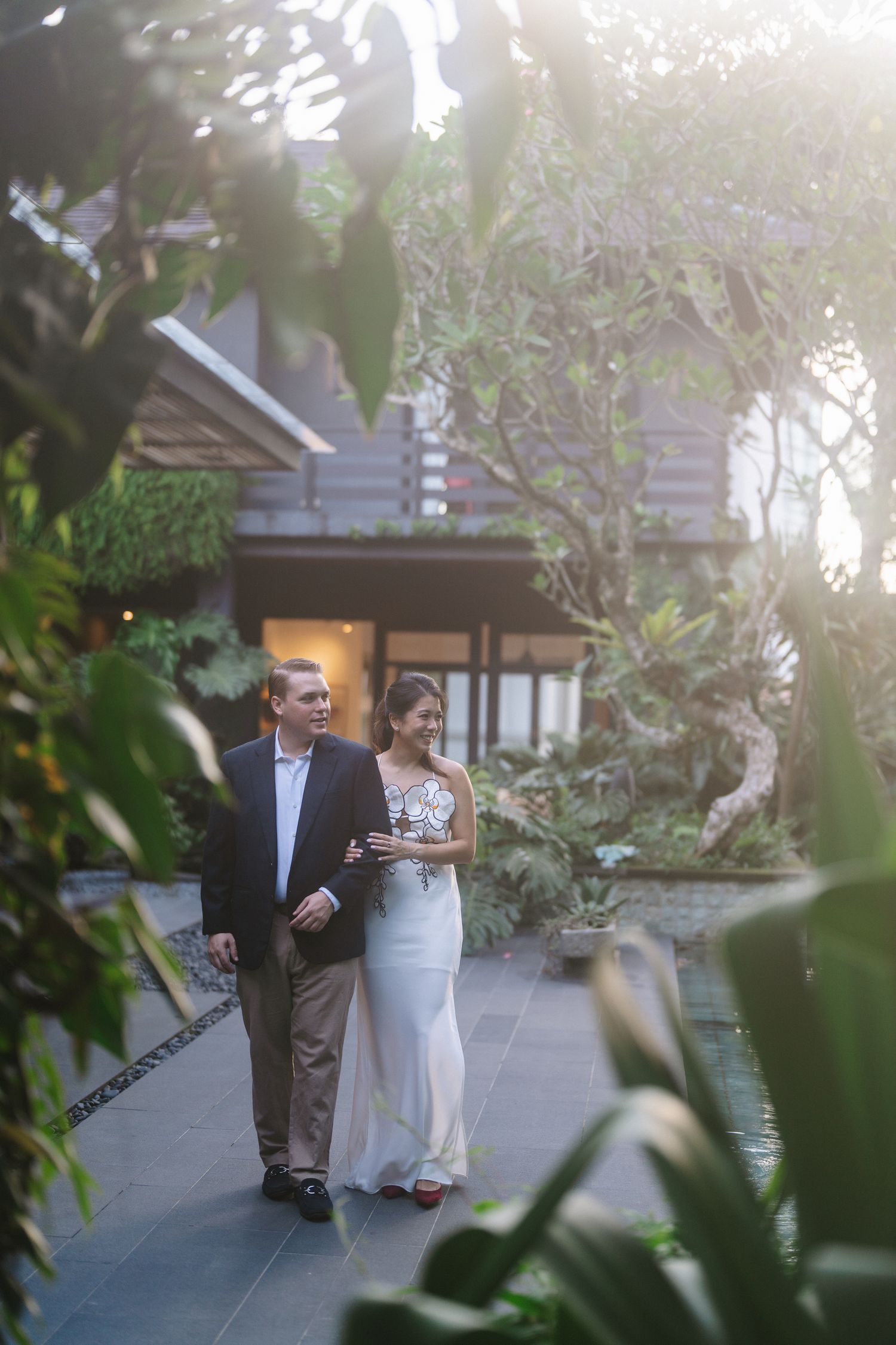 Couple in wedding attire walking through lush tropical garden pathway at sunset near elegant villa.