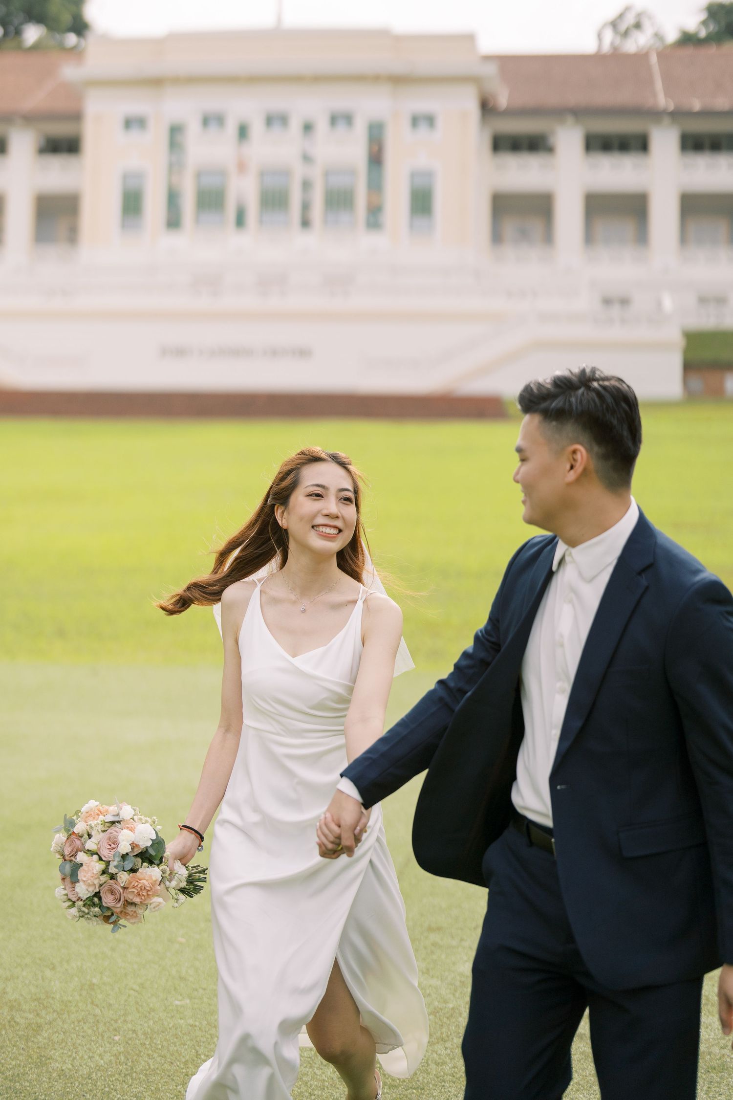 Newlyweds running playfully across a green lawn in formal attire.