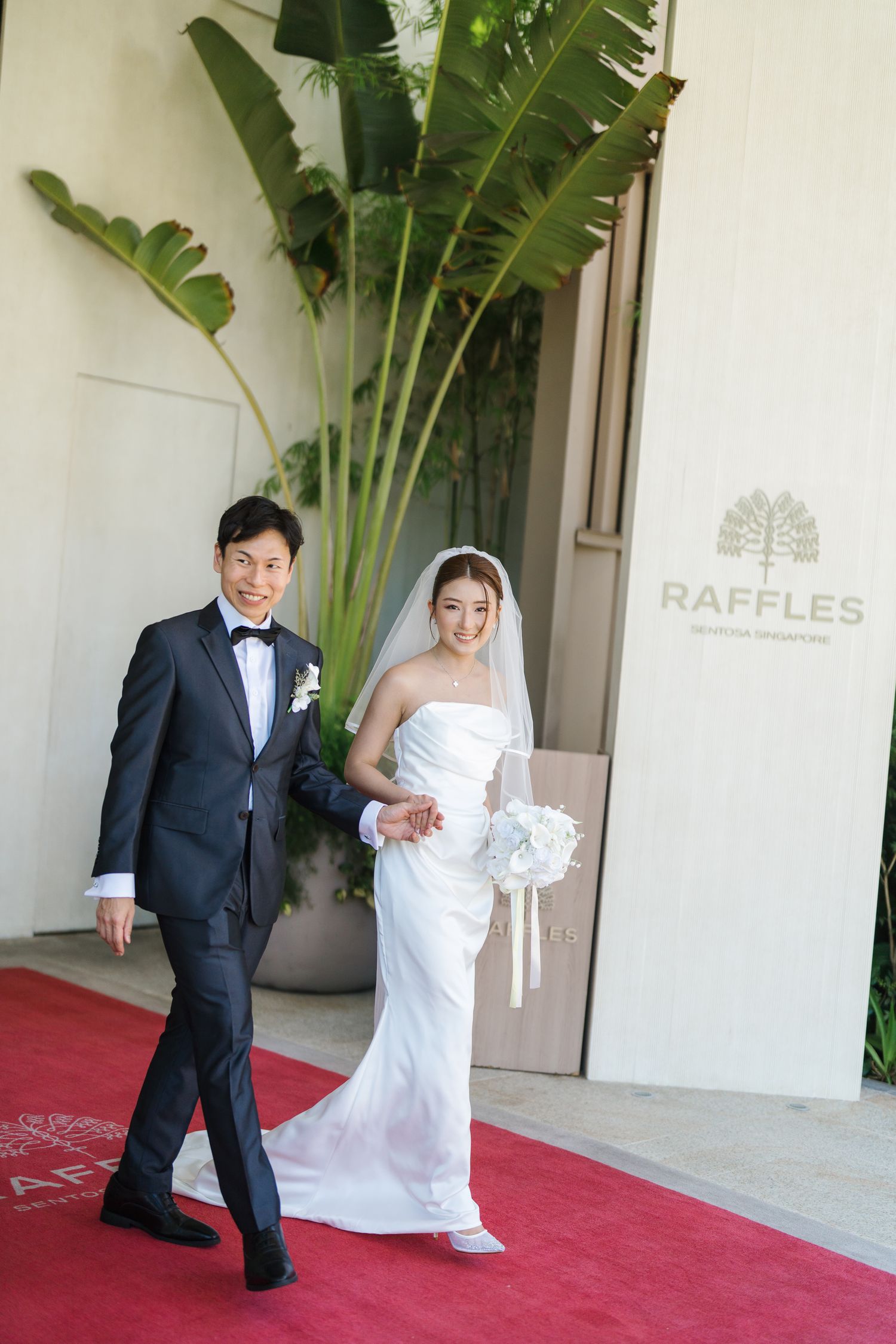 Bride and groom walk the red carpet at Raffles hotel, she in a white gown with veil, he in a dark tuxedo.