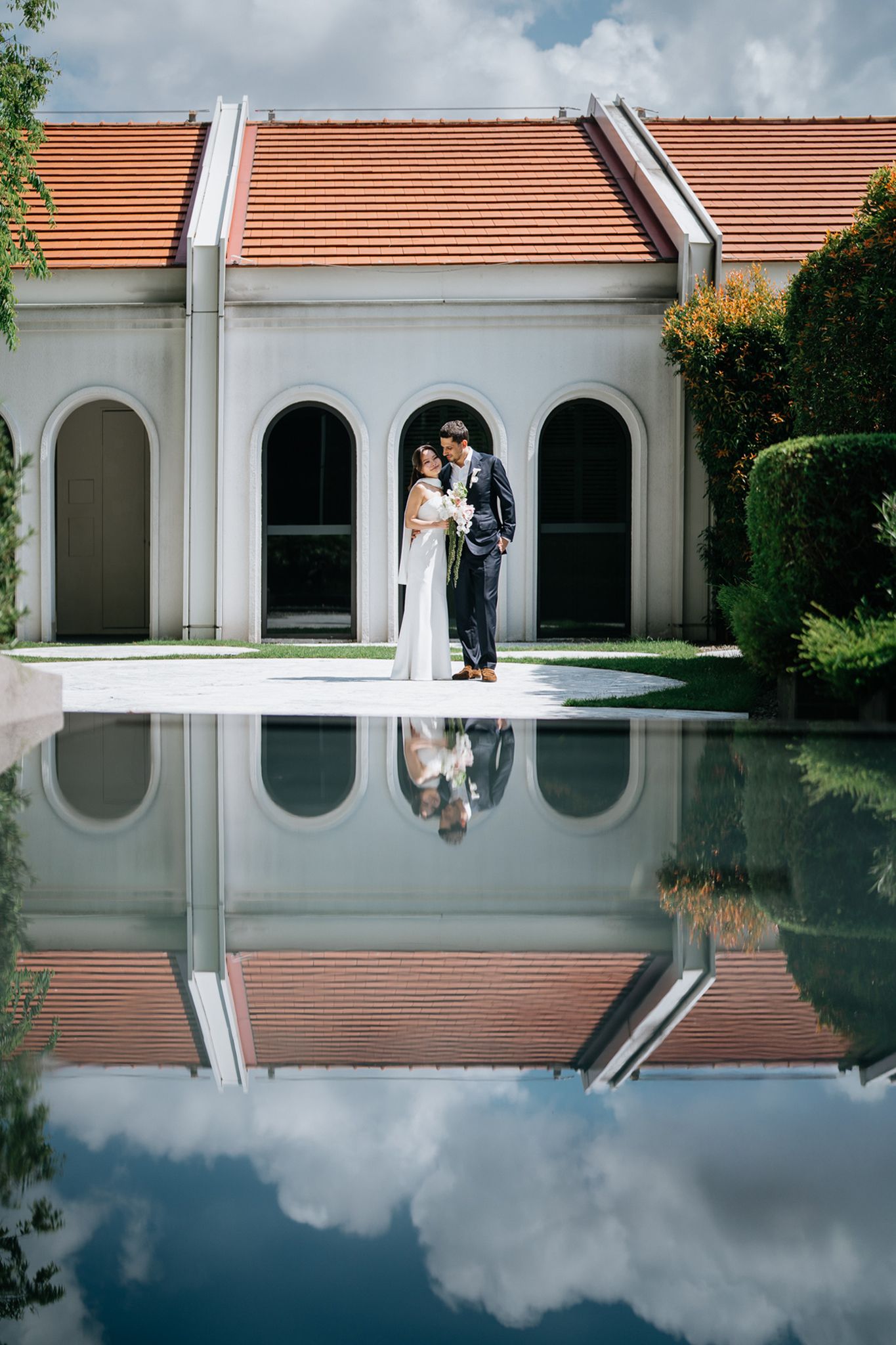 Bride and groom share a romantic moment beside a reflective pool at an elegant venue with arched windows.