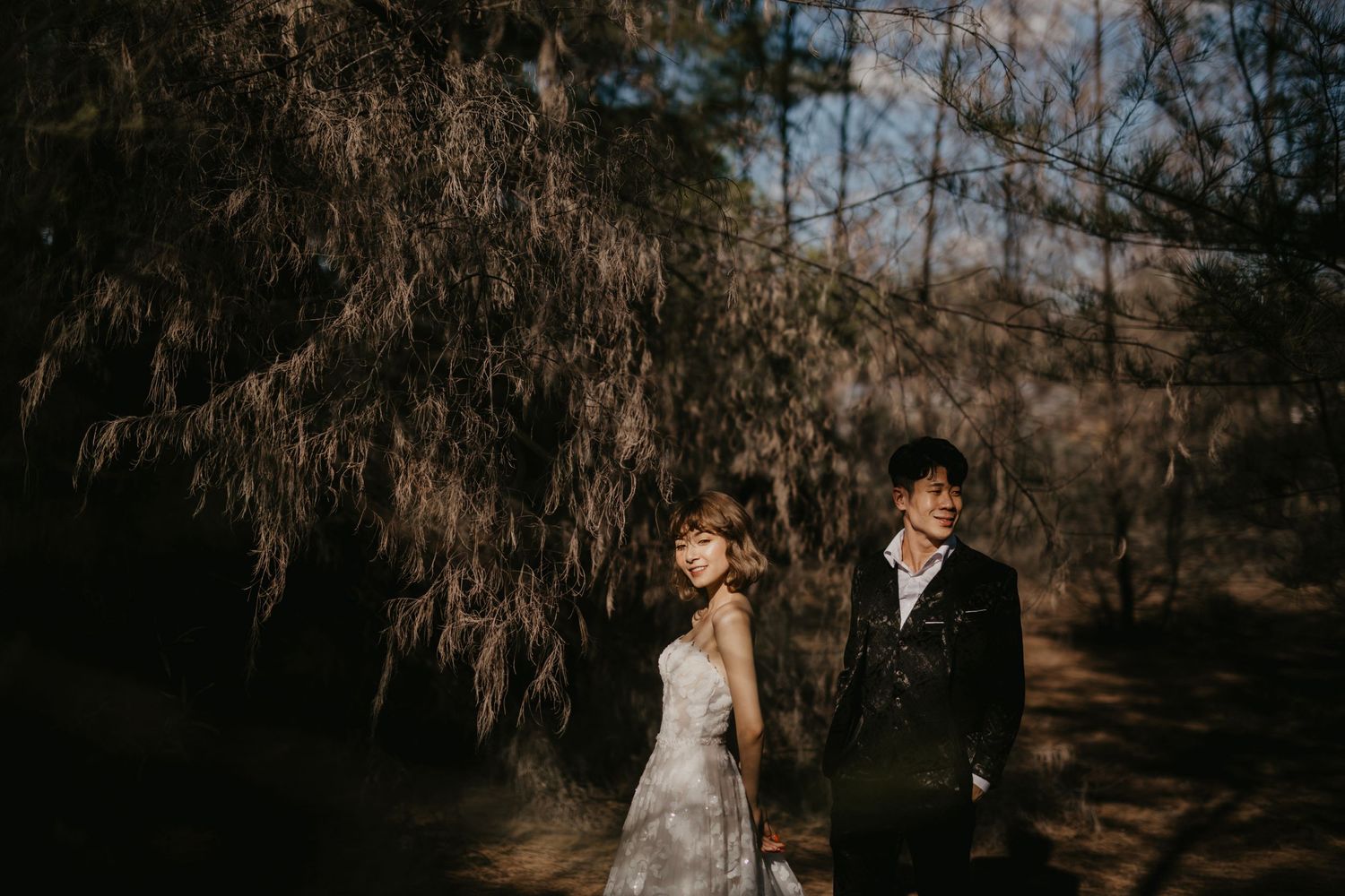 A wedding couple poses against dark trees during a moody evening photoshoot.