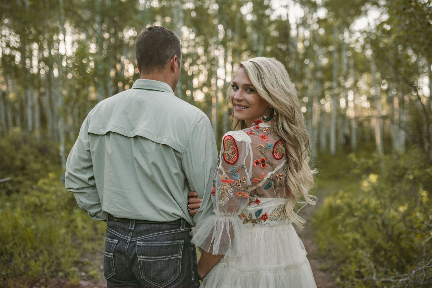 A couple standing together amongst green aspen trees and the woman is looking back at the camera, photographed by Kristy Baker Photography.
